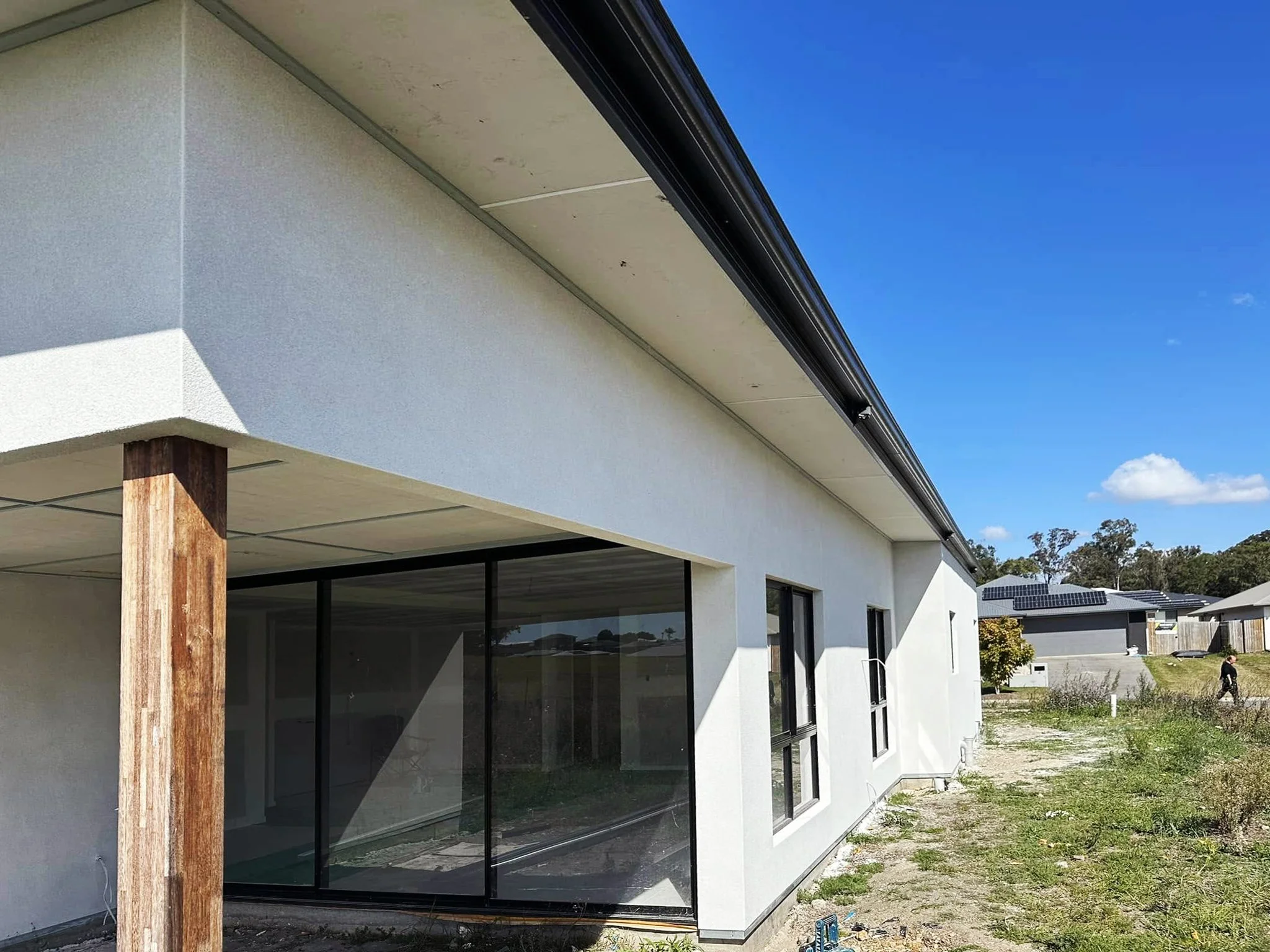 Modern house exterior featuring large glass windows, a flat roof, and vertical wooden support beam, with a clear blue sky in the background.