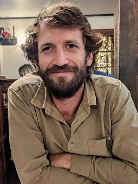 A man with a beard and wavy brown hair smiling, wearing a tan button-up shirt, sitting indoors with a rustic wooden wall and windows in the background.