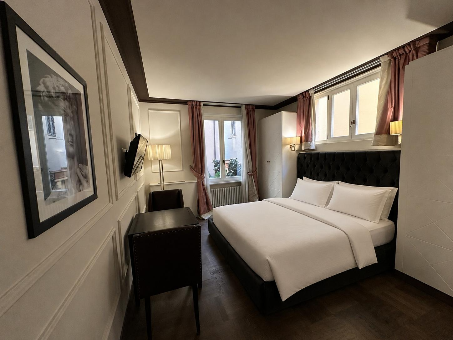 Hotel room with a king-sized bed, dark upholstered headboard, white bedding, pink curtains, wall-mounted lamps near the bed, a window on the side, a wall-mounted TV, a small table with a chair, and artwork on the wall.