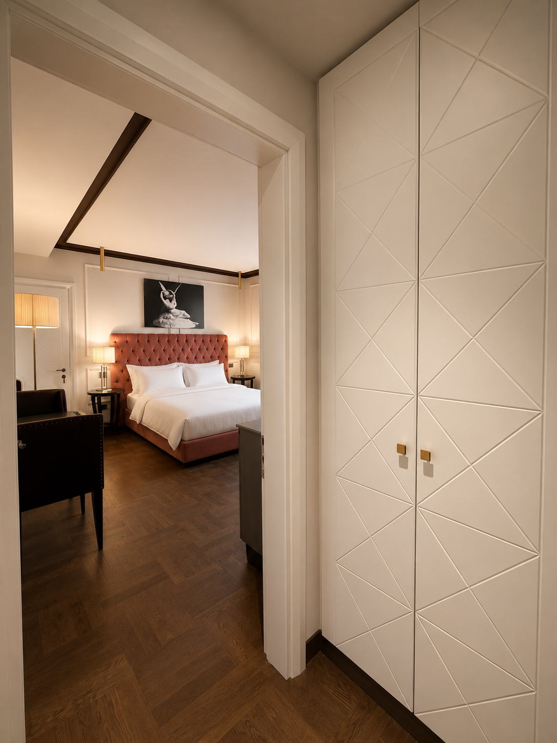 A view of a bedroom through a doorway, showing a bed with a pink quilted headboard, two bedside tables with lamps, a black and white artwork of a dog on the wall, a black chair, a floor lamp, and a dark wooden floor.