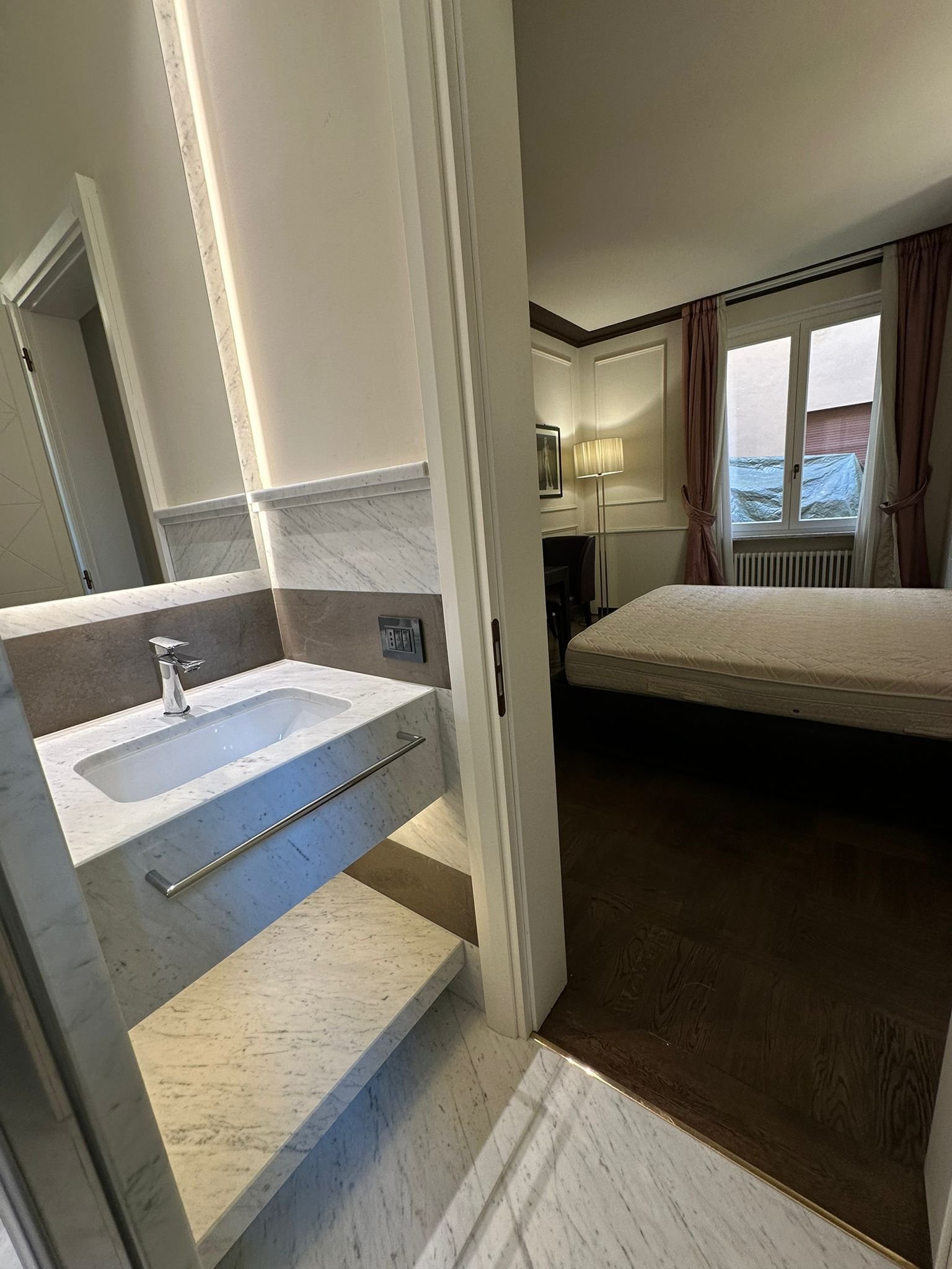 View of a hotel room with a bed near the window, a lamp, a framed picture, and a small desk. The bathroom with a sink and marble counter is visible through an open door.