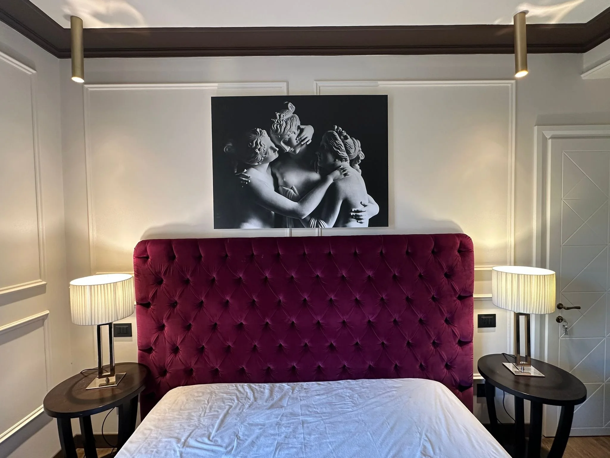 A bedroom with a velvet, button-tufted, deep pink headboard, a white bedspread, black side tables with lamps, a black and white sculpture artwork on the wall, cream-colored walls with decorative paneling, a white door with geometric patterns, and mod