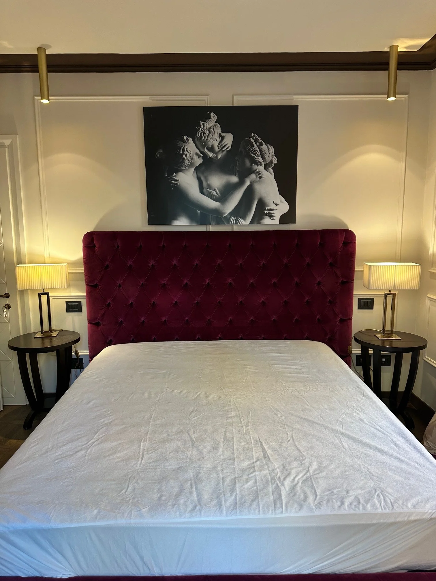 A bedroom with a bed, two bedside tables with lamps, a large dark red headboard, and a black-and-white painting of three people hugging on the wall.