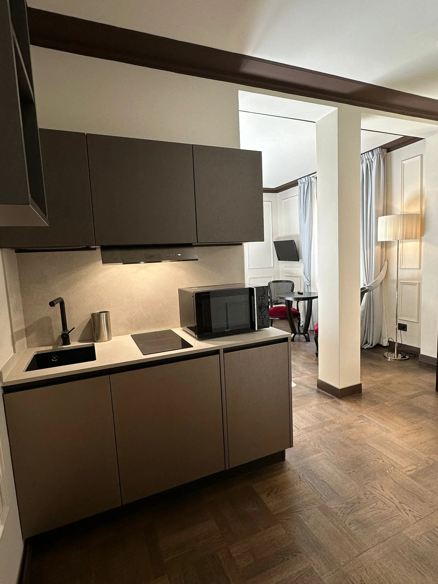 A kitchen area with gray cabinets, a black microwave, a small round table with a red chair, a floor lamp, and windows with light-colored curtains.