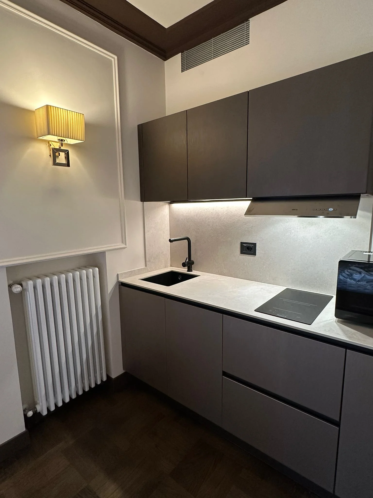 Modern kitchen with dark wood cabinets, beige countertops, black sink, black stovetop, stainless steel range hood, and white wall with decorative molding and a lamp, radiator, and electrical outlet.