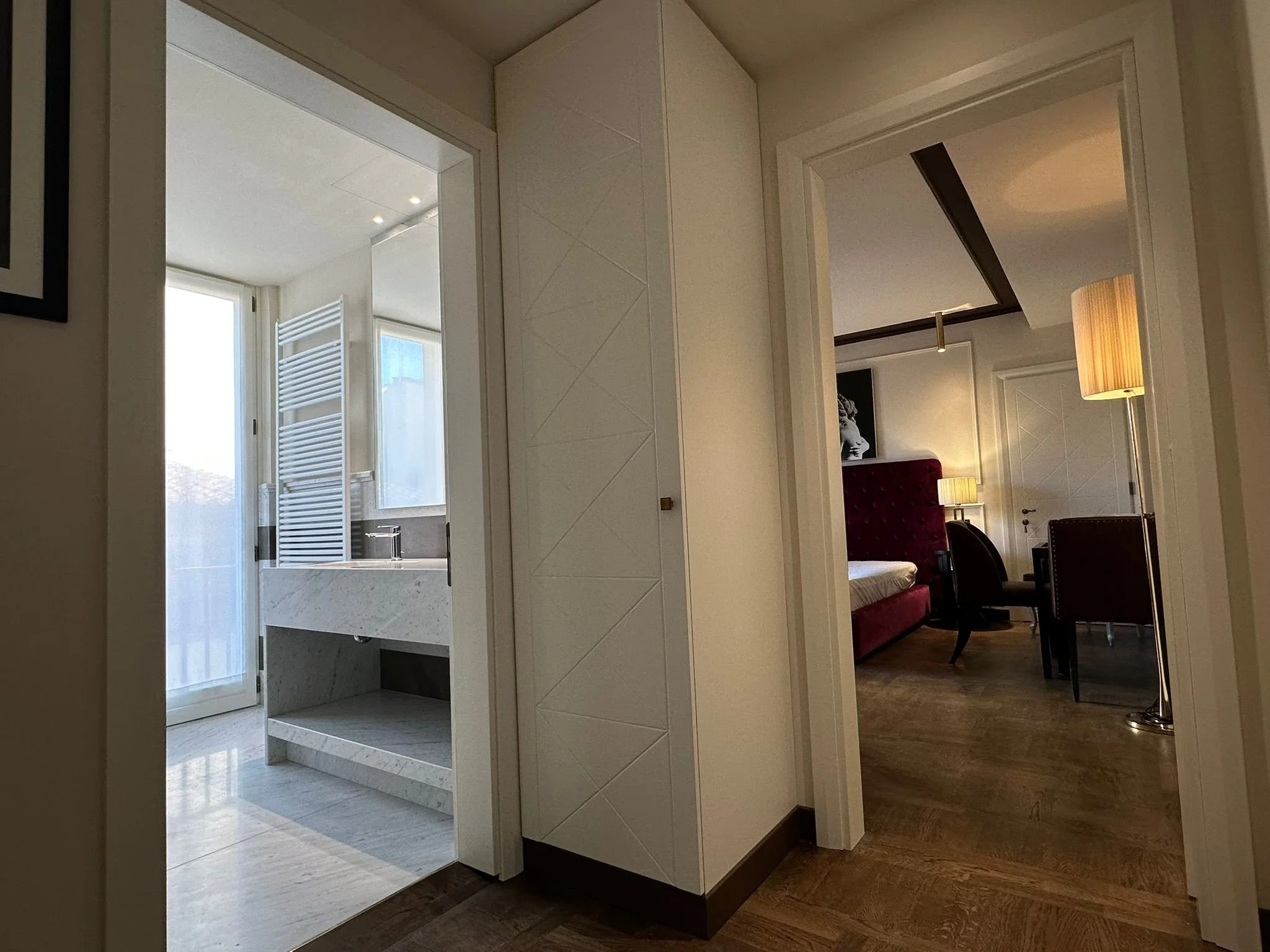 Interior view of a room with a white partition wall, a staircase with a glass railing leading to a balcony, and a sitting area with a red velvet sofa and floor lamps.
