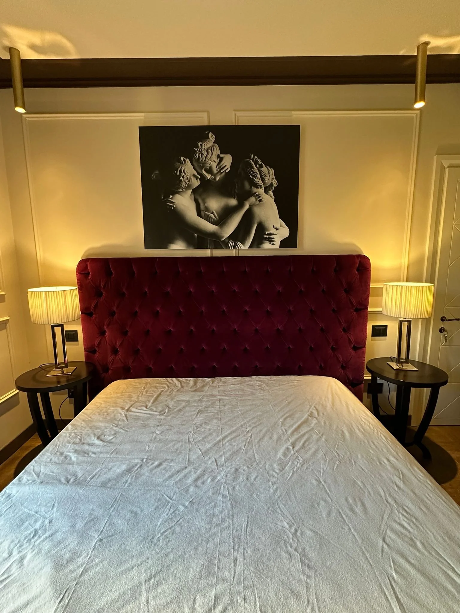 A bedroom with a burgundy tufted headboard, two black bedside tables with modern lamps, and a large black-and-white painting of three embracing classical sculptures hanging on the beige wall.