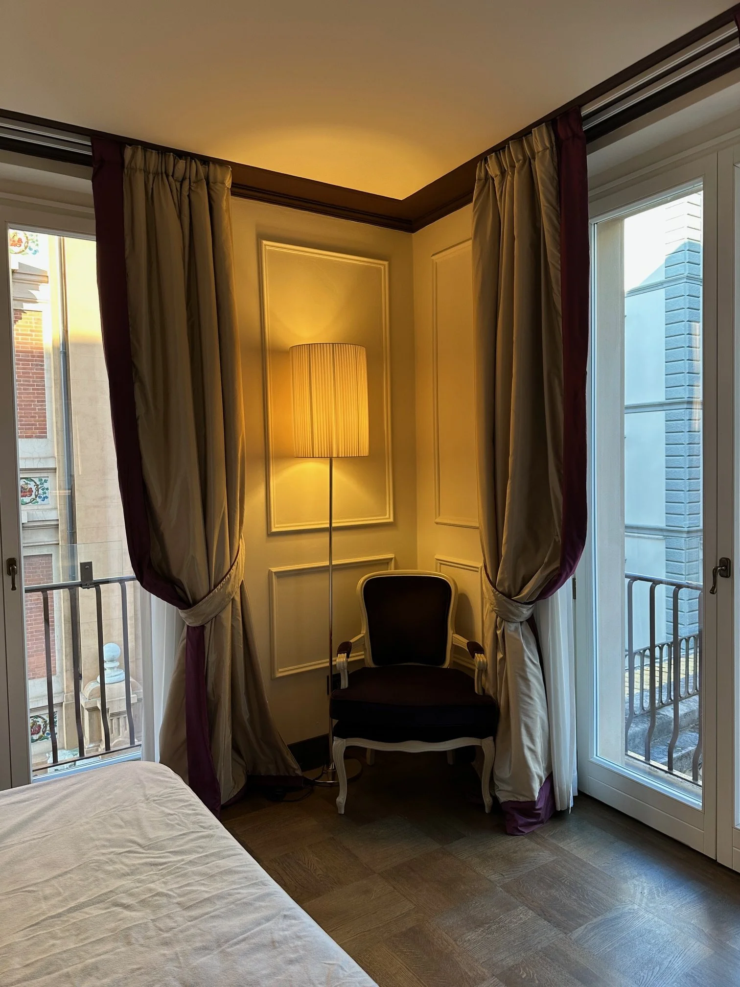 A cozy corner of a room with large windows and curtains, a chair, and a tall lamp casting warm light.