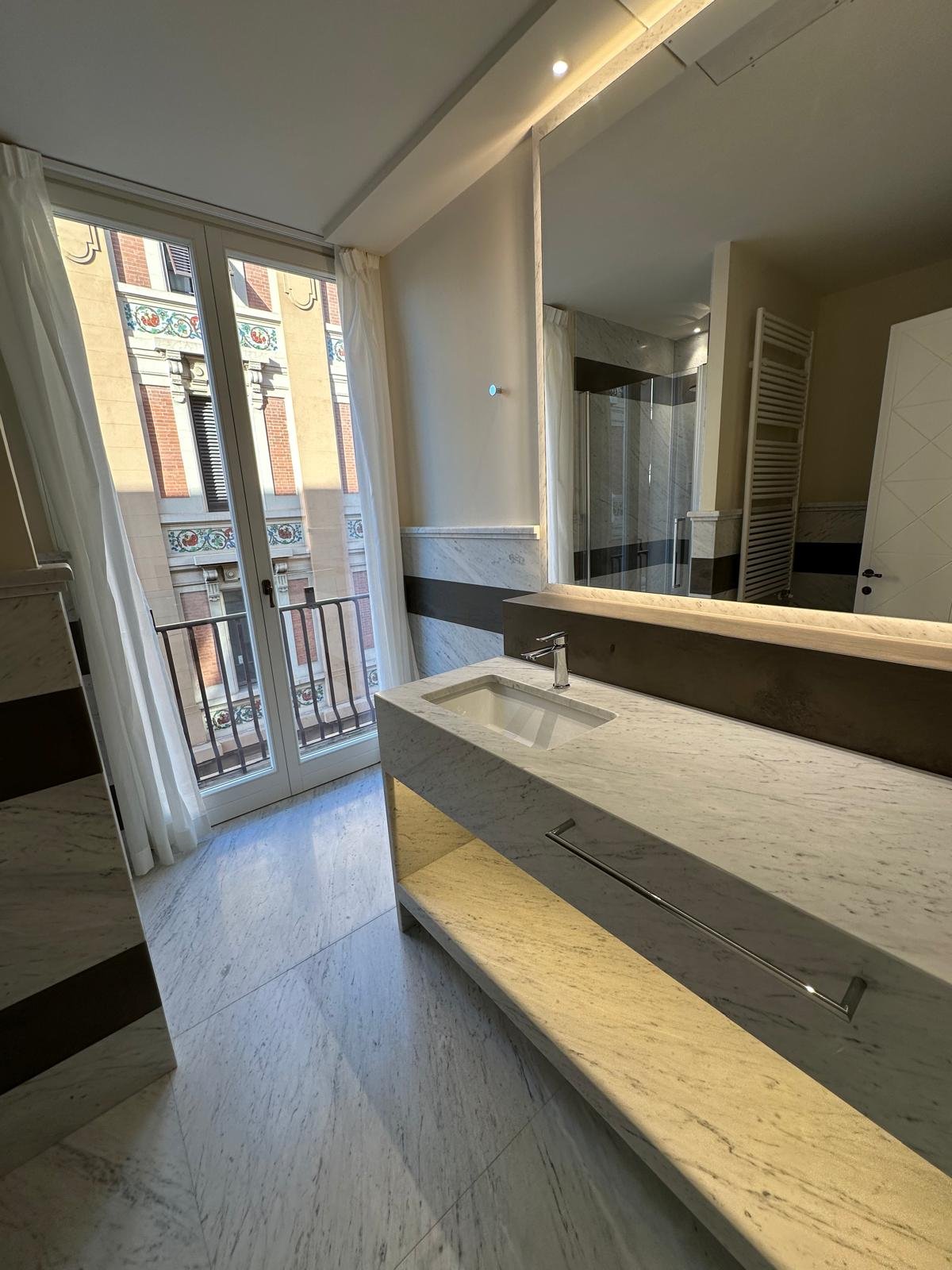 Modern bathroom with marble countertop, large mirror, and glass-enclosed shower, adjacent to a window with a view of a historic building.