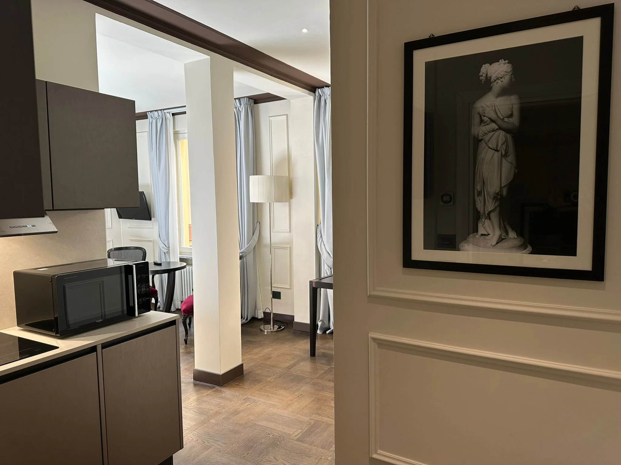 A modern, open-concept living space with a kitchen area visible in the foreground, featuring a microwave and a stovetop. The background includes a dining area with a window dressed with white curtains, and a living room with a lamp and seating. A fra