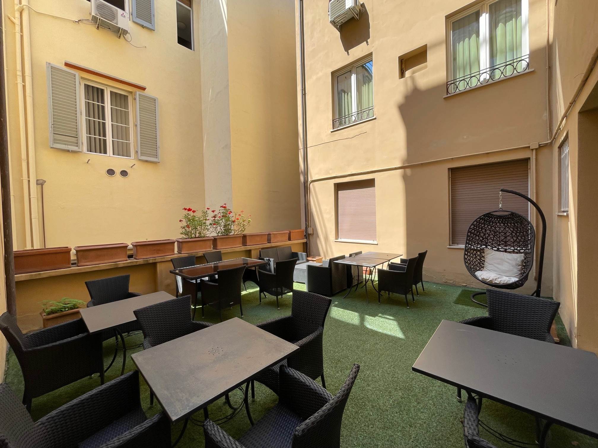 Apartment courtyard with outdoor furniture, potted plants, and hanging chair, surrounded by beige and cream-colored walls with windows and air conditioning units.