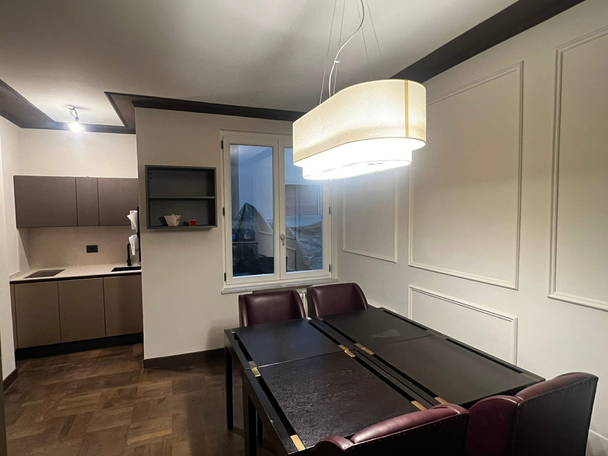 A dining room with a black table and maroon chairs. There's a large modern ceiling light fixture, a window, and a small shelf with items on it. The room has decorative wall panels and dark trim along the ceiling.