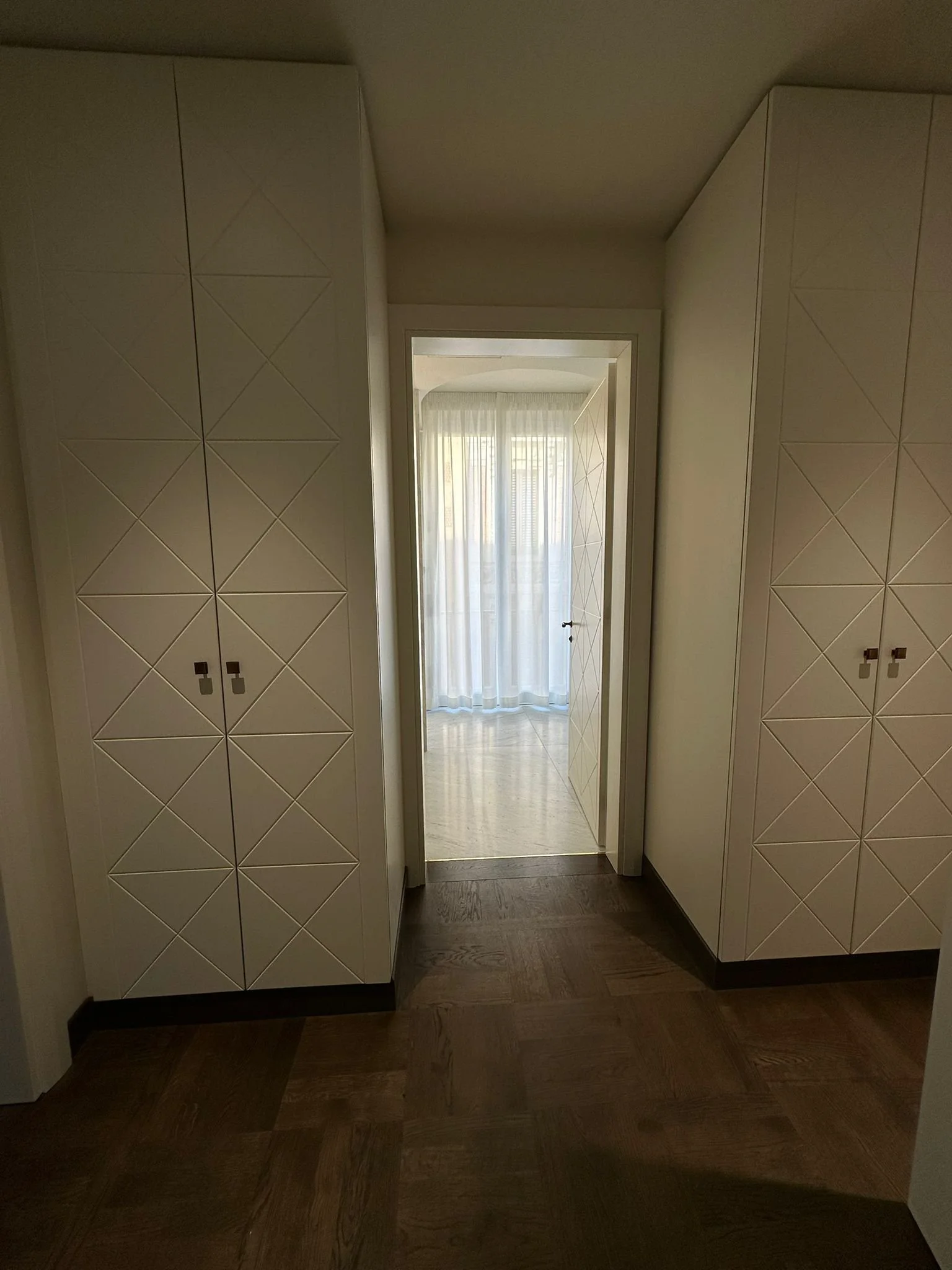 A hallway with built-in white cabinets on both sides, leading to a room with sheer curtains and natural light, dark wooden flooring.