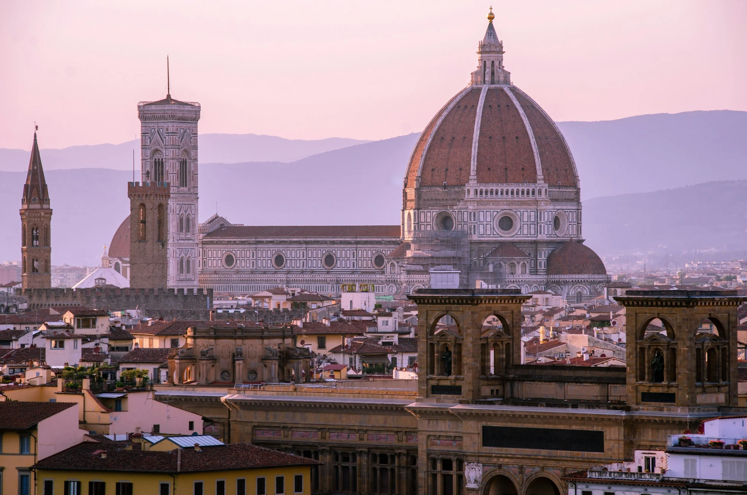 View of Florence, Italy with the Florence Cathedral (Duomo) featuring a large red-tiled dome, surrounded by historic buildings and mountains in the background.