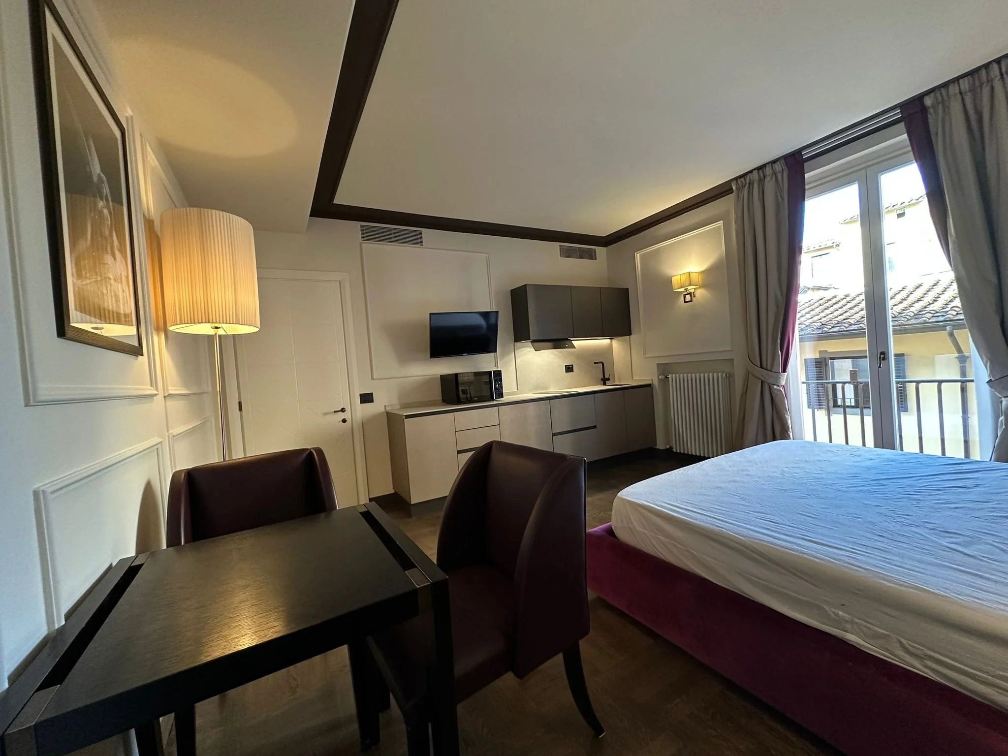A hotel room with a bed, a dining table with chairs, a wall-mounted TV, kitchen cabinets, large window with curtains, and wall lamps.