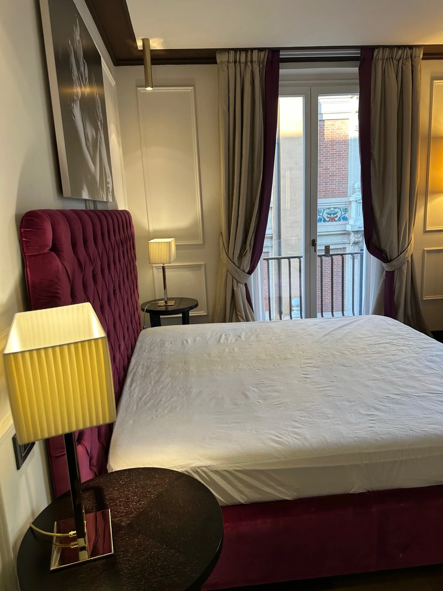 Interior of a hotel room with a bed, a side table with a lamp, a purple upholstered headboard, curtains covering a balcony door, and a photograph on the wall.