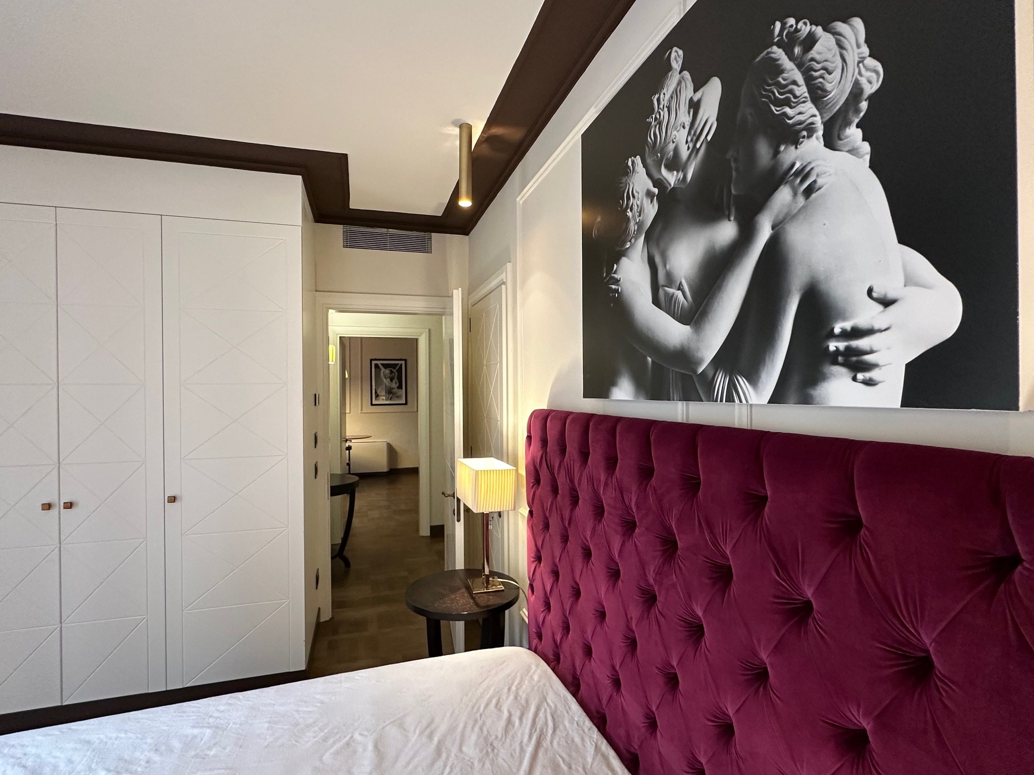 Hotel room with white wardrobe, maroon tufted headboard, black and white wall sculpture of a family, side table with lamp, and doorway to hallway.