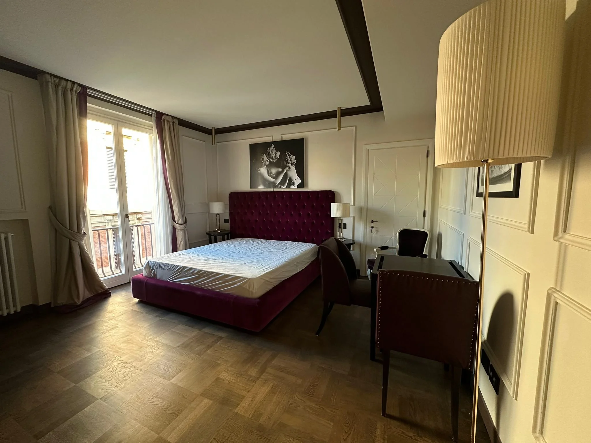 Interior of a hotel room with a large windows, cream curtains, a red upholstered headboard, a bed with white sheets, and various lamps and furniture.