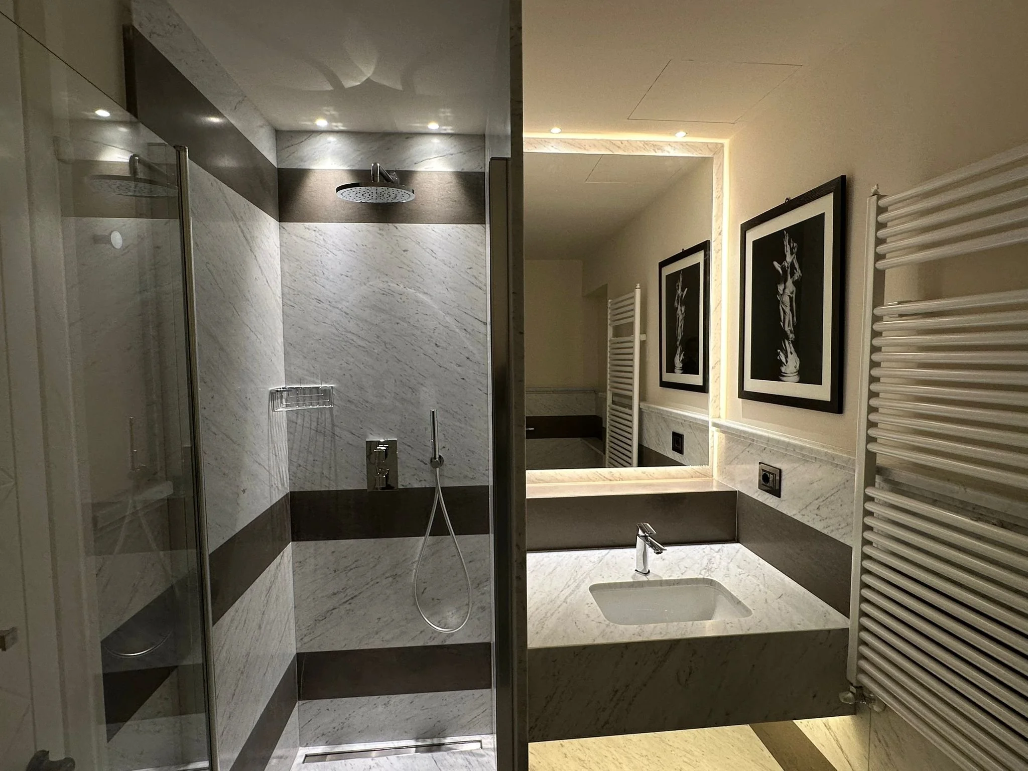 A modern bathroom with a shower area featuring marble walls and a rain showerhead, a white sink with a chrome faucet, and framed black-and-white artwork on the wall. There is also a heated towel rail and ambient lighting.