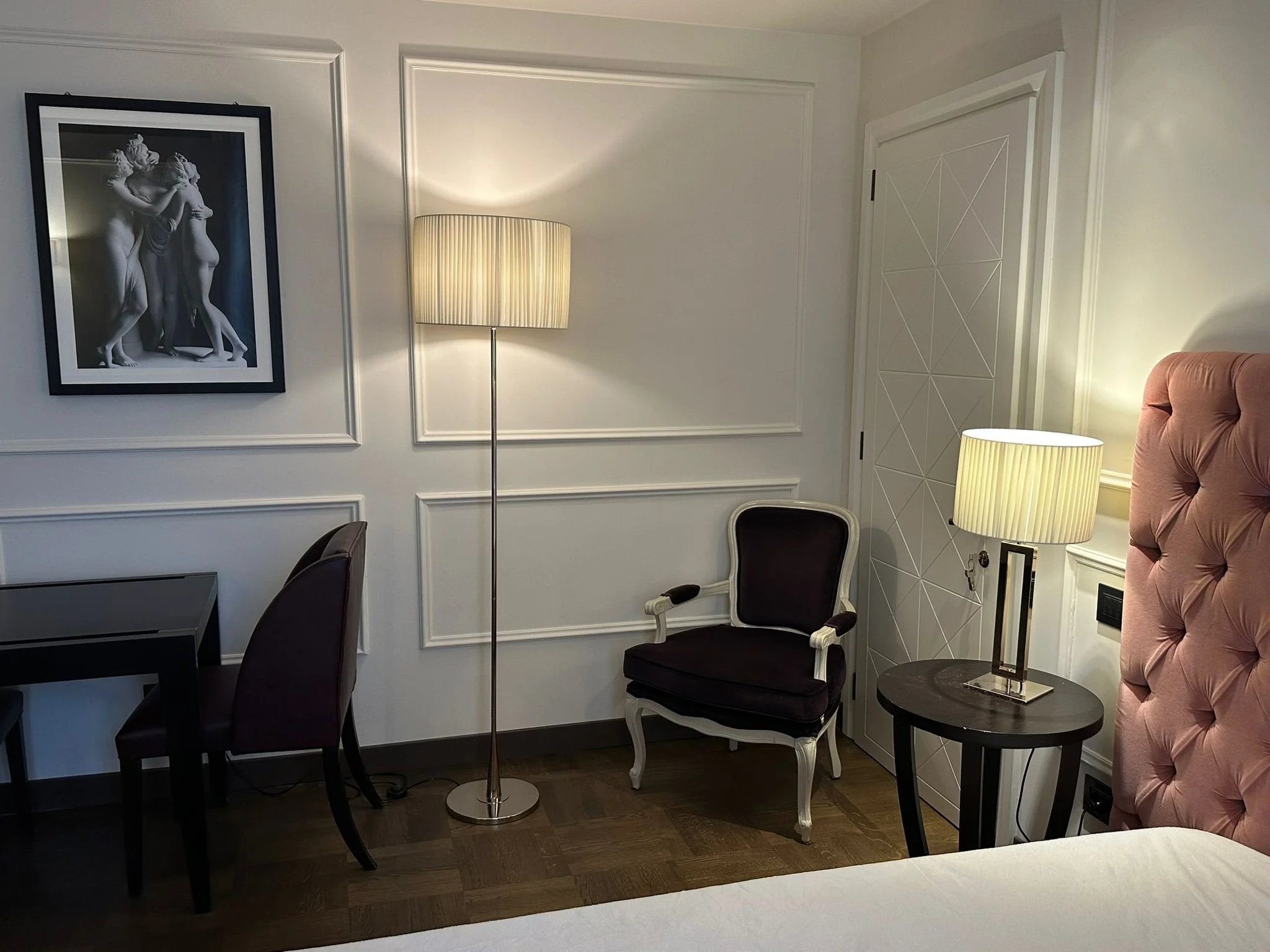 Interior of a hotel room corner showing white paneled walls, two standing lamps with cream shades, a black side table, two chairs, and a pink tufted armchair. A black and white framed artwork of three Classical-style figures is on the wall.