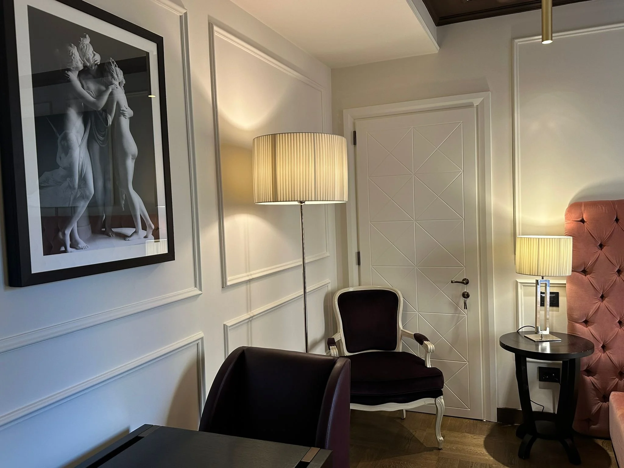 Interior room with framed black and white sculpture photograph on the wall, two table lamps, white paneled door, and a pink tufted chair.