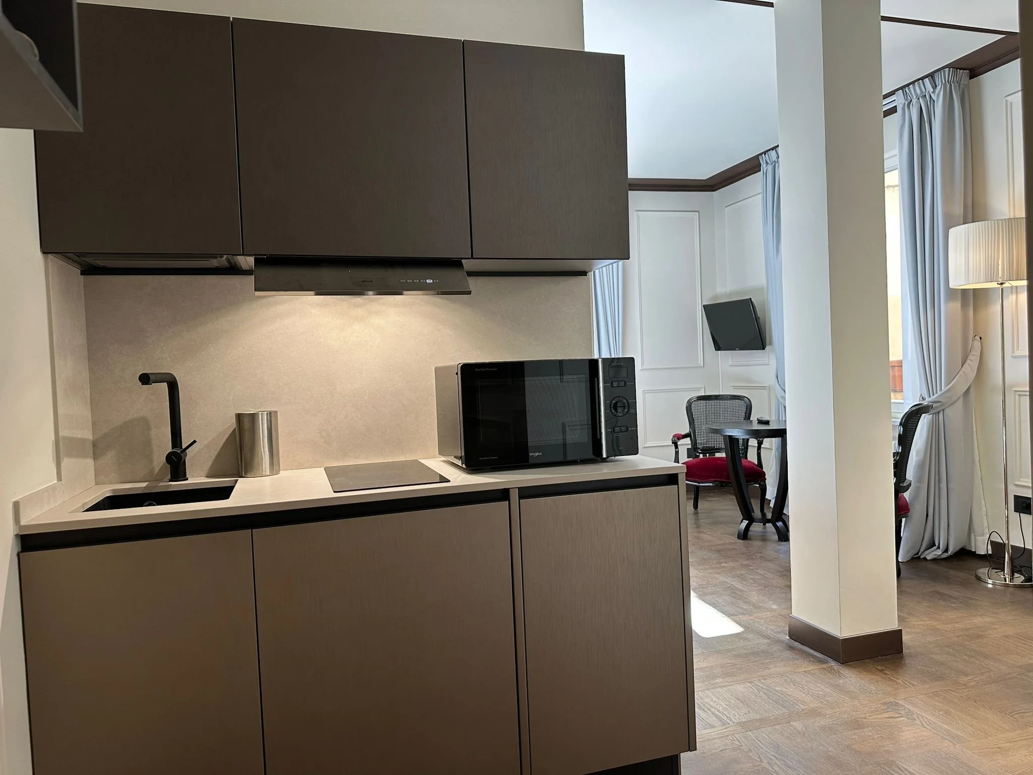 A modern, open-concept kitchen with dark upper cabinets, a white countertop with a black sink and faucet, and a microwave on the counter. In the background, there are large windows with light curtains, a small dining table with chairs, and a standing