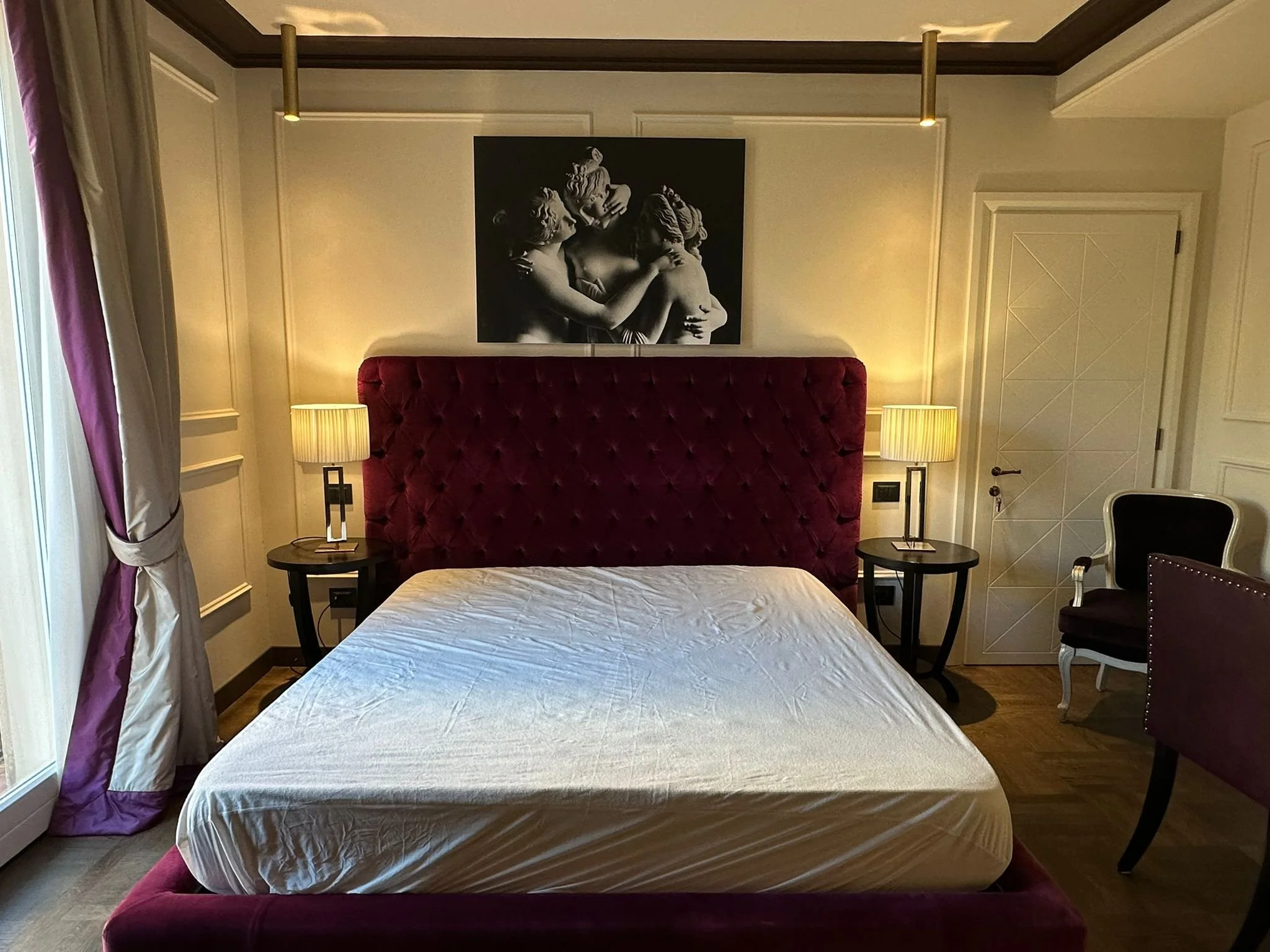 Hotel bedroom with purple upholstered headboard, two bedside tables with table lamps, a black and white wall art, and a window with purple curtains.