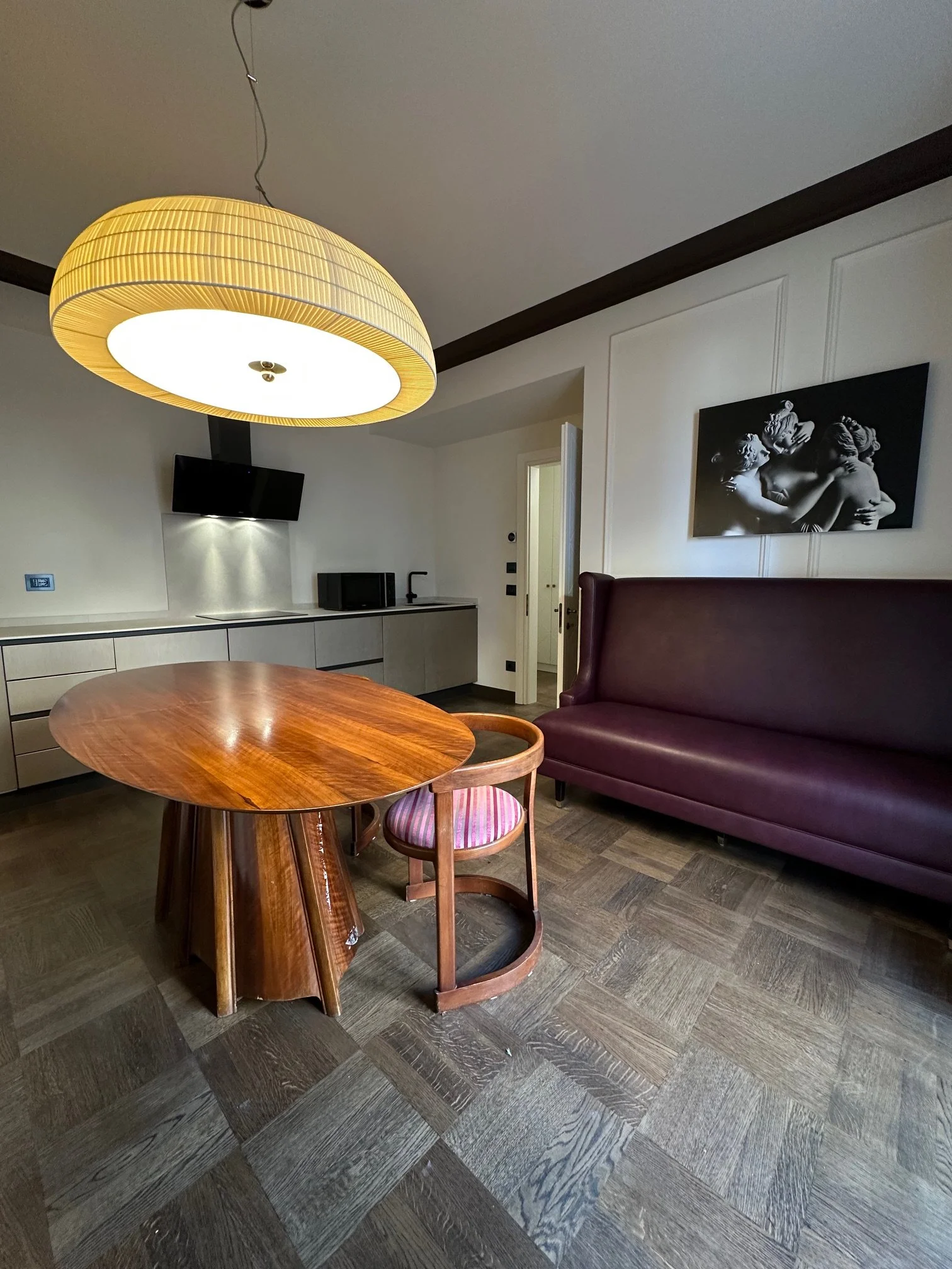 Living room with a round wooden dining table, a pink velvet sofa, wall art of a sculpture, a mounted flat-screen TV, and a large overhead yellow lamp with a white light
