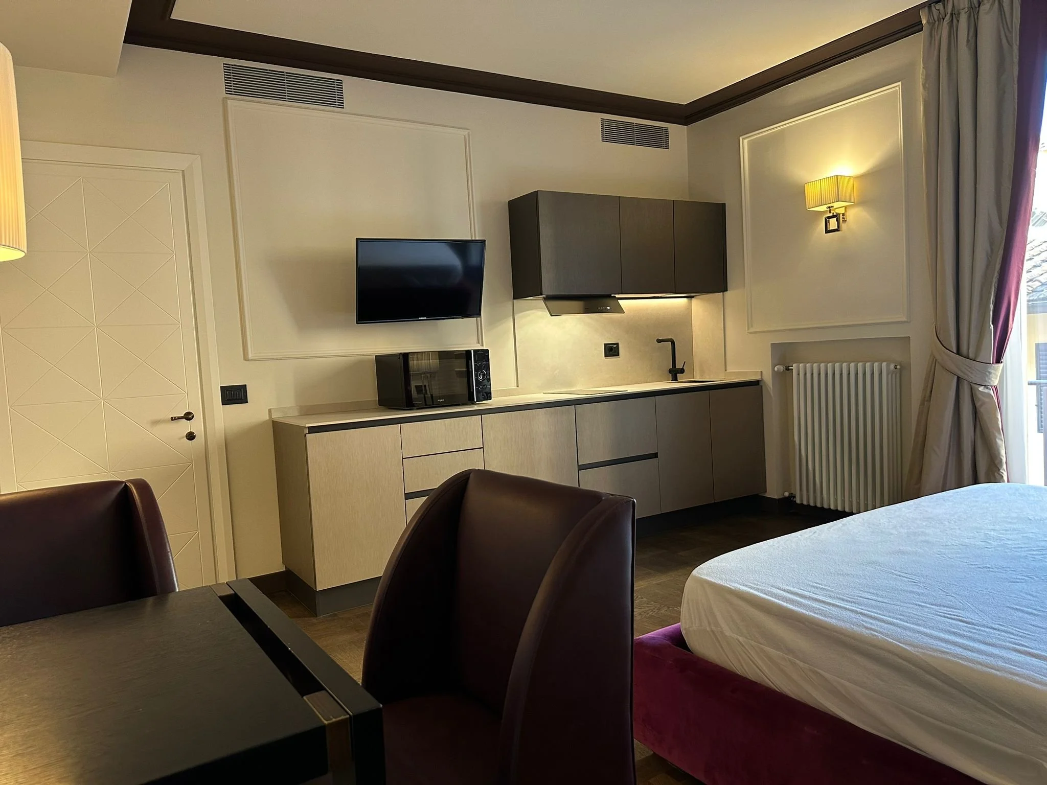 Modern hotel room with a bed, dark wood chairs, a small black table, a kitchenette area with a microwave and cabinets, a wall-mounted TV, and a window with curtains.