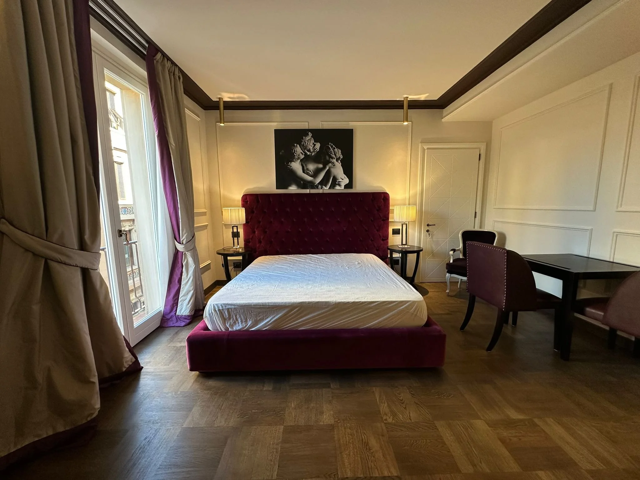 A bedroom with a large bed featuring a red upholstered headboard, flanked by two side tables with lamps. There are large windows with curtains on the left side, and a black and white artwork hanging above the headboard. A desk with a purple chair is 