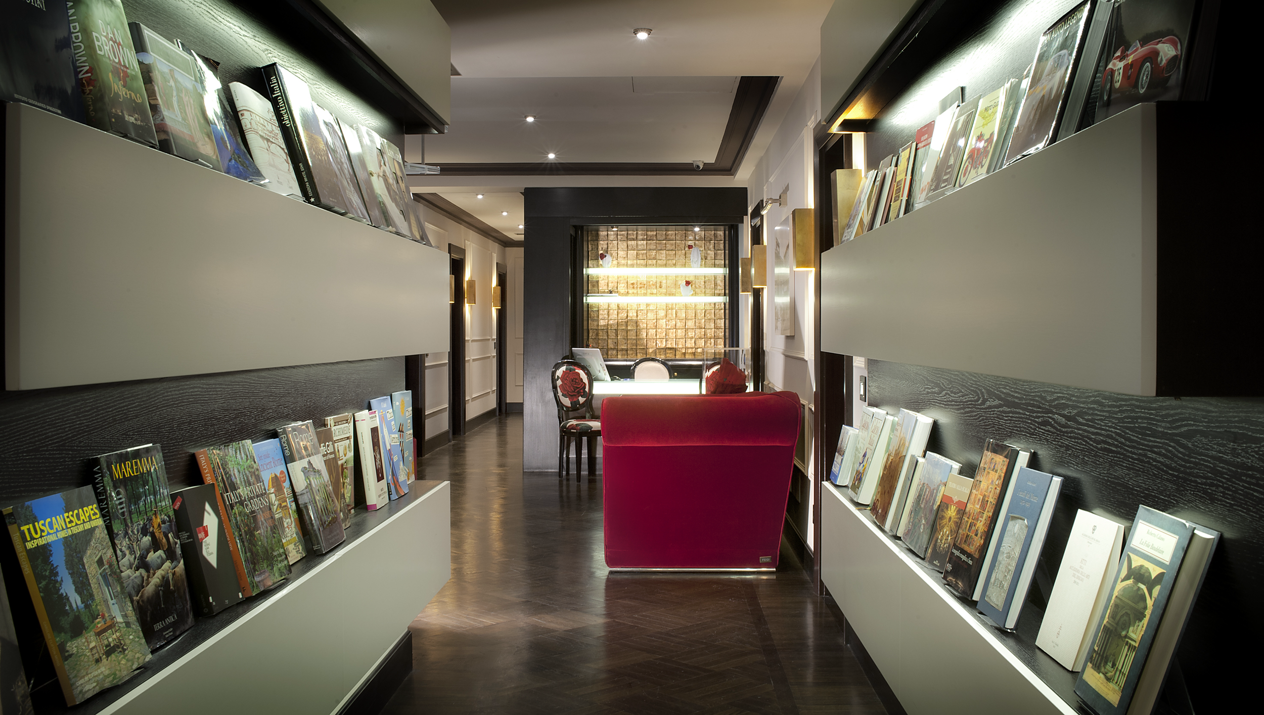 Interior of a bookstore with shelves of colorful books on both sides, a red armchair in the center, and a window at the back with decorative artwork and seating area.