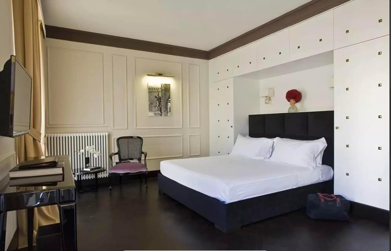 A bedroom with a tufted pink headboard, two small side tables with lamps, a black and white painting of a dancer on the wall, and a window with curtains showing rooftops outside.