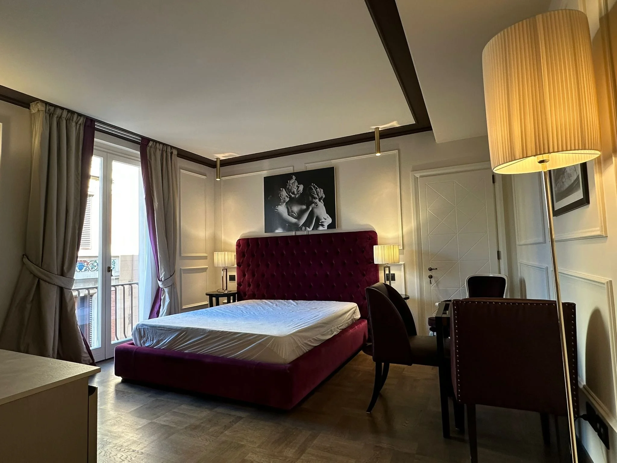 Hotel room with a large bed with a tall, red velvet headboard, two bedside lamps, a framed black and white photo of sculpture, a black chair, dining table, and floor-to-ceiling windows with curtains.