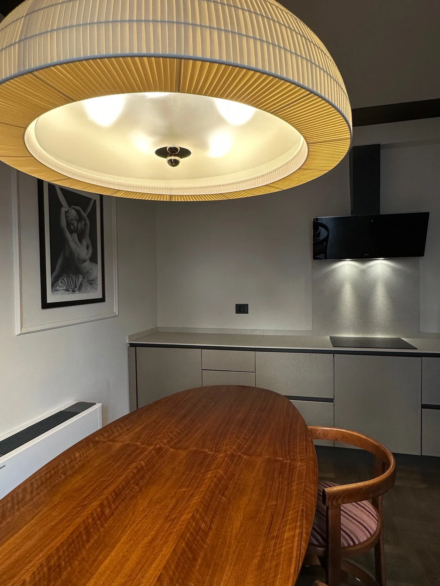 A room with a large wooden dining table and a wooden chair with striped cushion, a modern ceiling light, wall art of a black-and-white photograph of a woman, a mounted flat-screen TV, and a white cabinet.