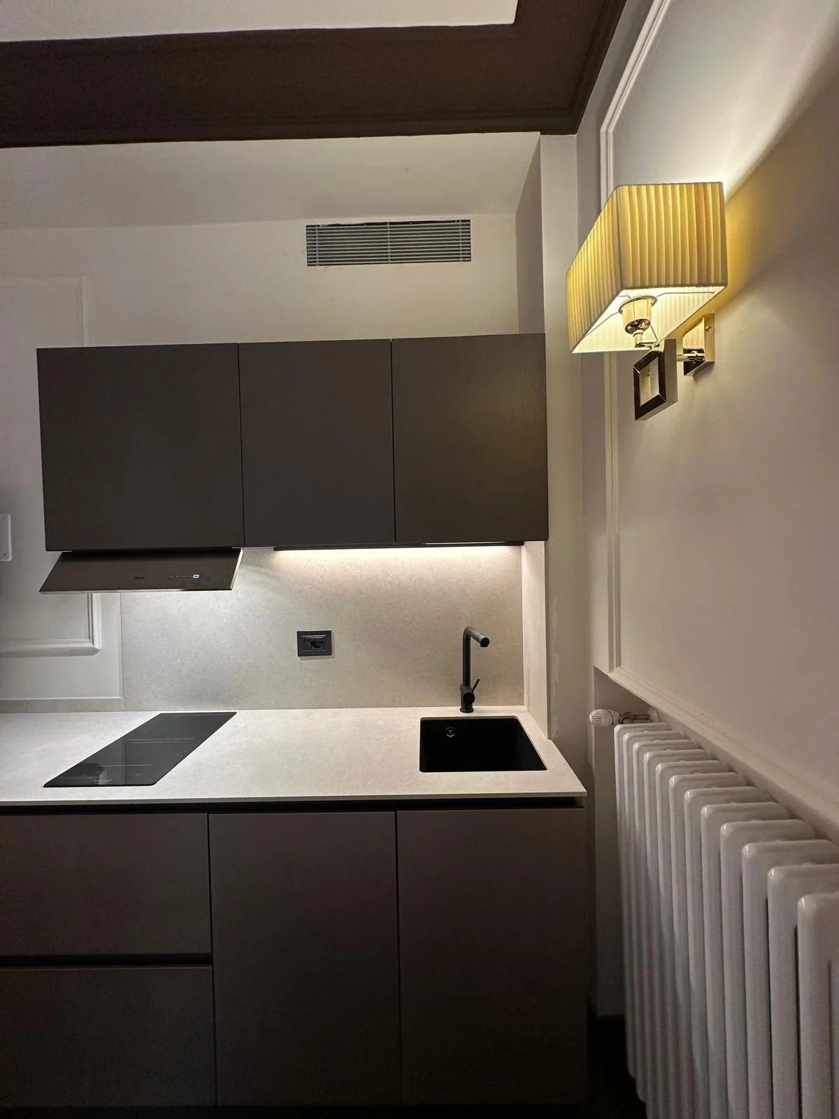 Modern kitchen with gray cabinets, black sink, black stovetop, and a beige wall-mounted lamp.