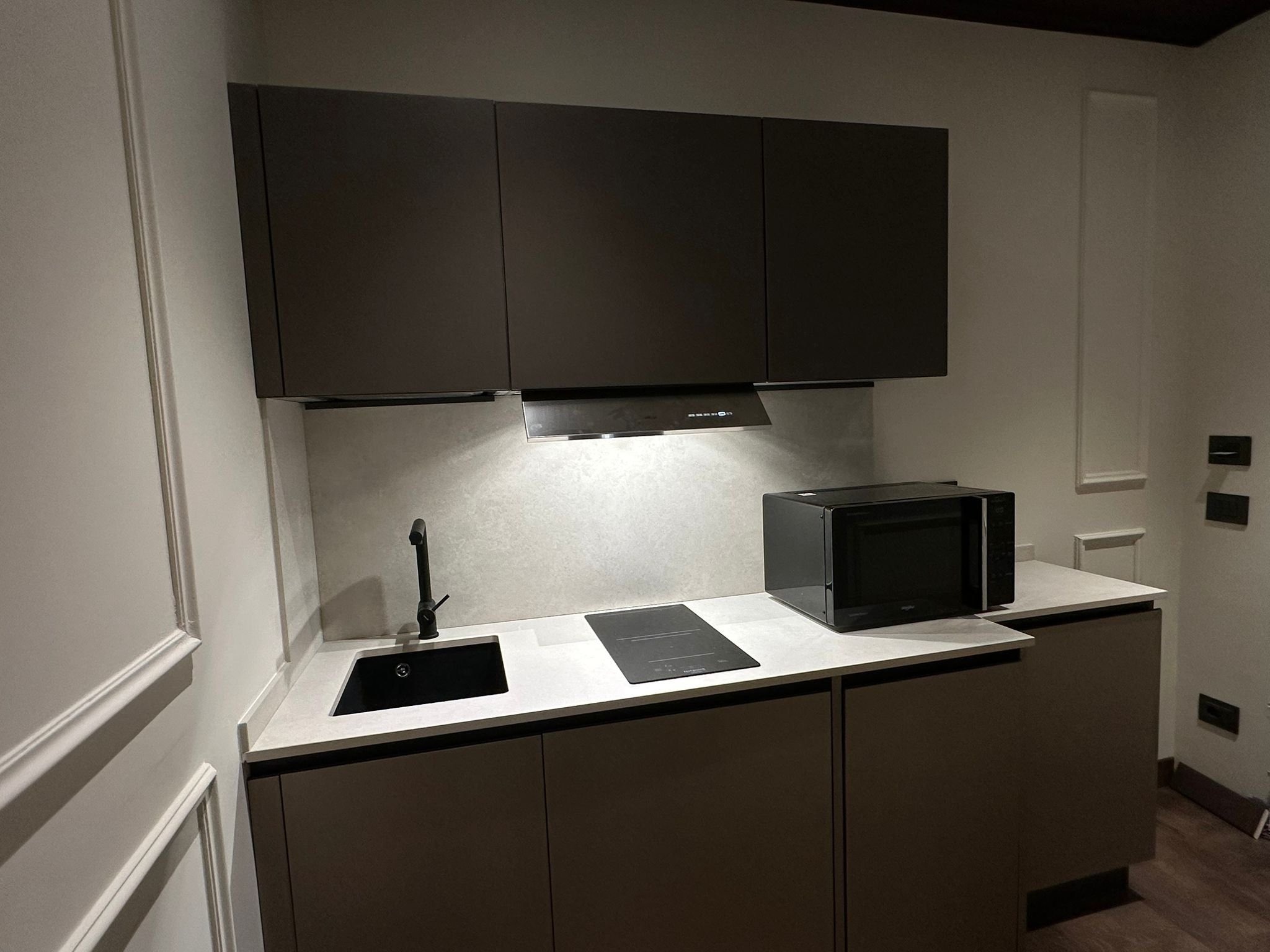 Modern kitchen with dark cabinets, black stove, microwave, white countertop, and black sink, with white walls and wood flooring.