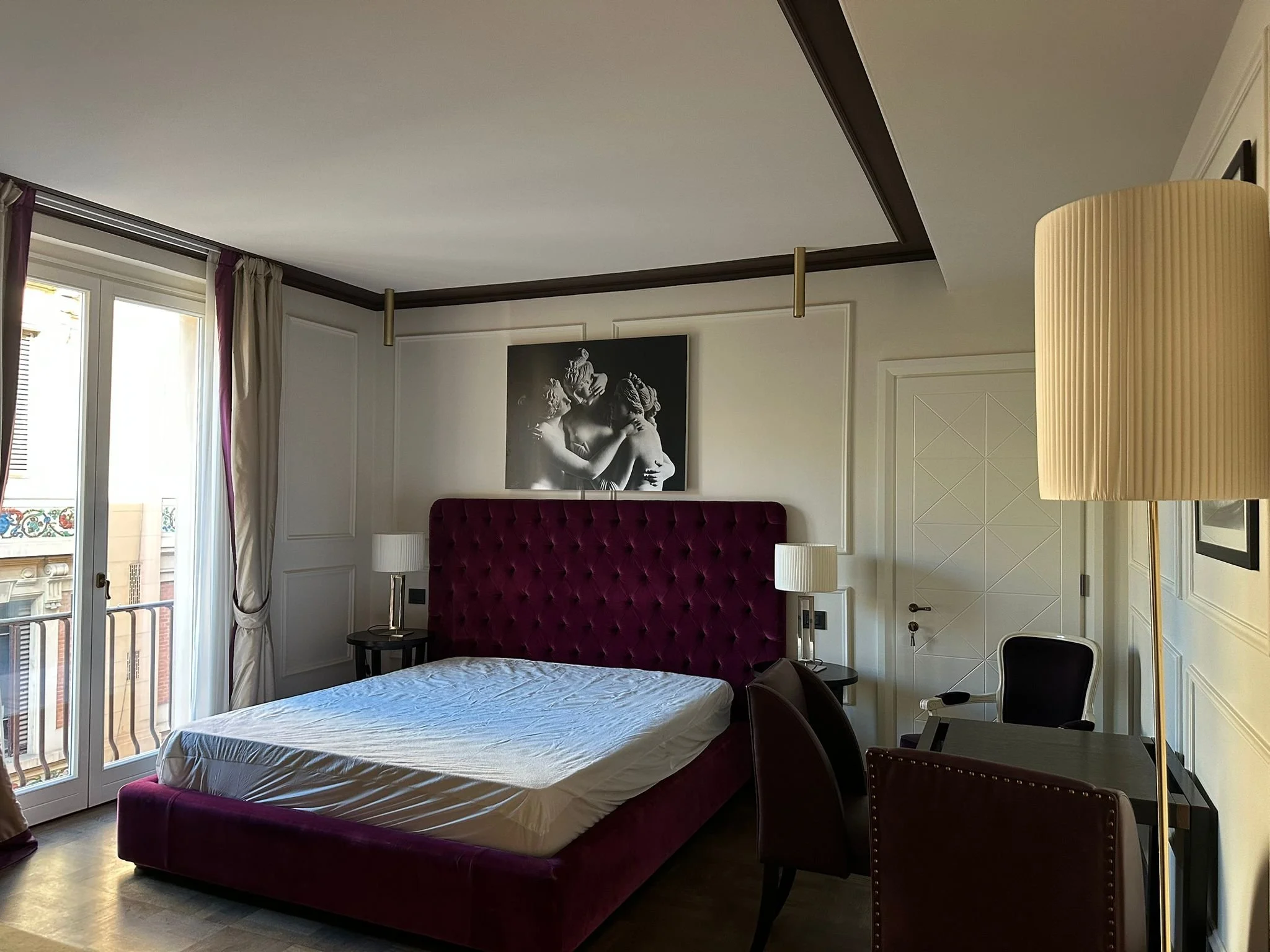 Interior of a bedroom with a bed with a velvet headboard, a nightstand, a picture of a sculpture of lovers, and a floor lamp. Large windows with curtains, balcony and city views.
