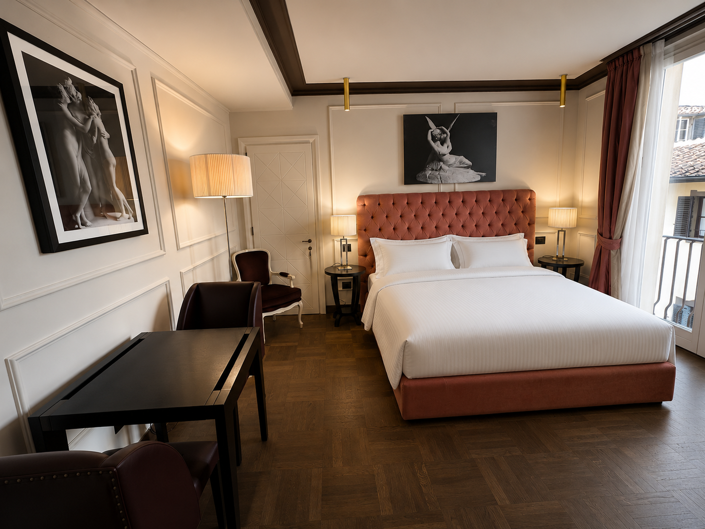 Hotel room with a large bed with pink tufted headboard, two side tables with lamps, artworks on white paneled walls, a wooden desk, and a glass door leading to a balcony with curtains.