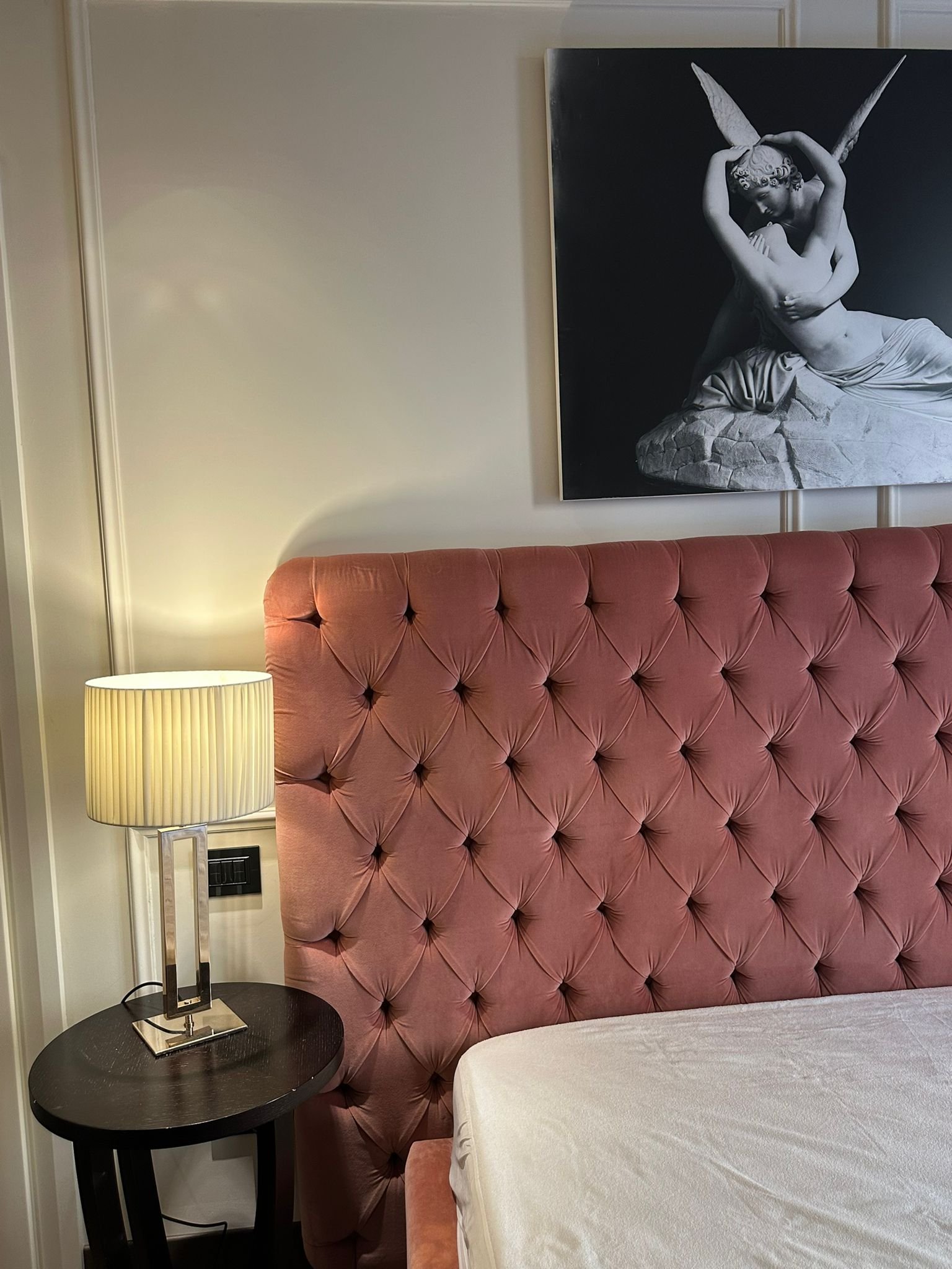 A pink tufted headboard behind a white bed sheet, with a table and lamp on the left side of the bed. A large black and white artwork of a woman with wings is hanging on the wall above the headboard.