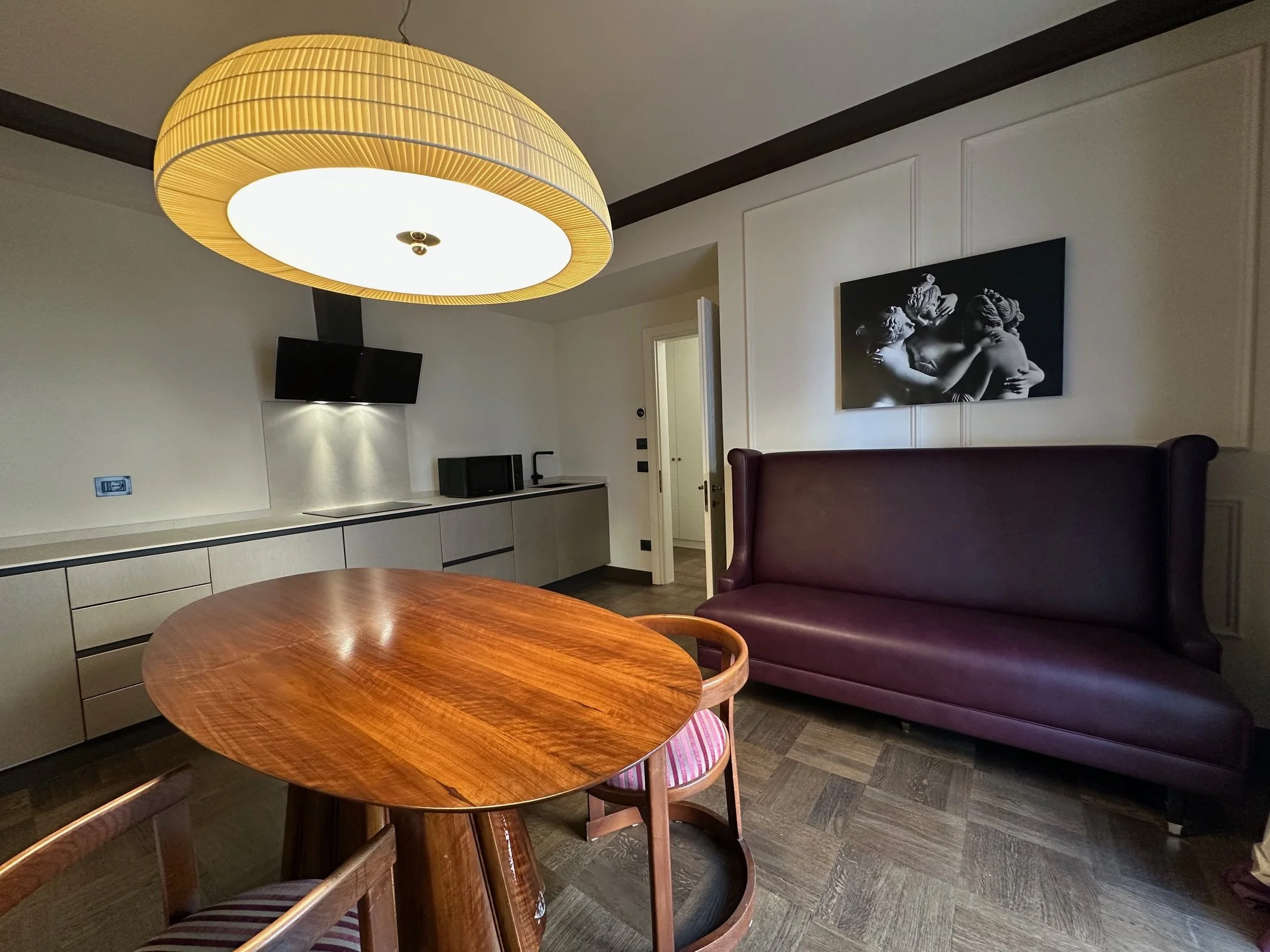 Living room with a large ceiling light, a black and white art piece on the wall, a maroon sofa, a circular wooden table, kitchen cabinets with a microwave, a door, a black TV, and a small striped chair.