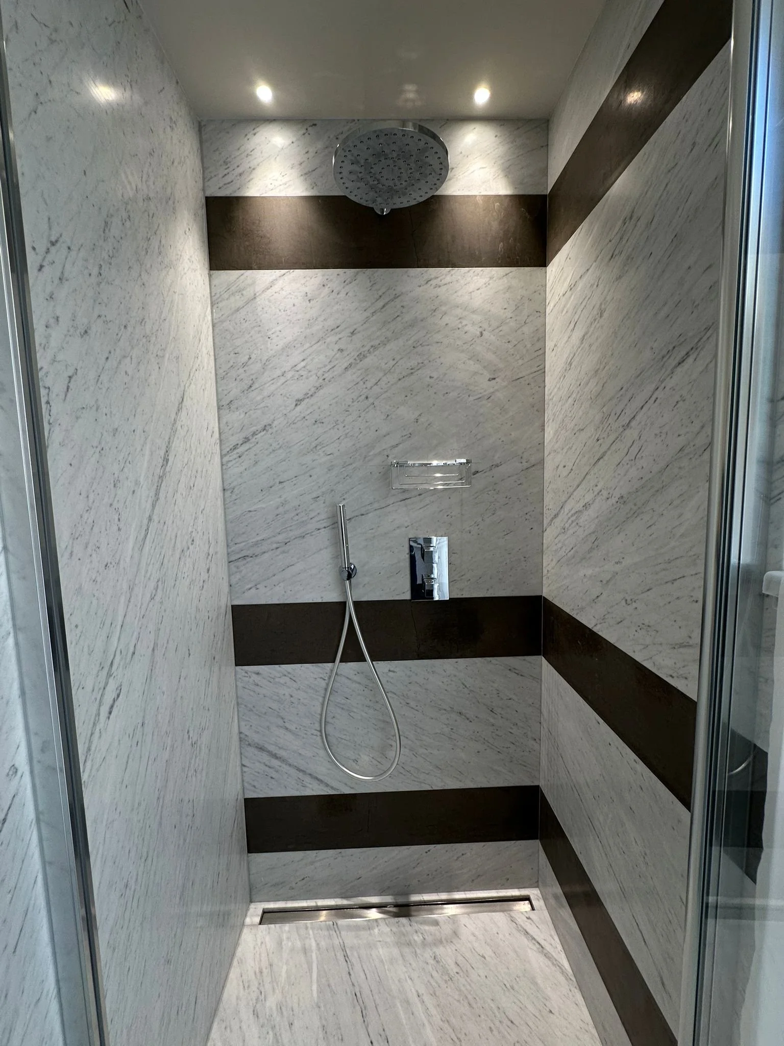 A modern shower with marble walls and floors, a round overhead showerhead, a handheld shower, and a linear drain at the base.