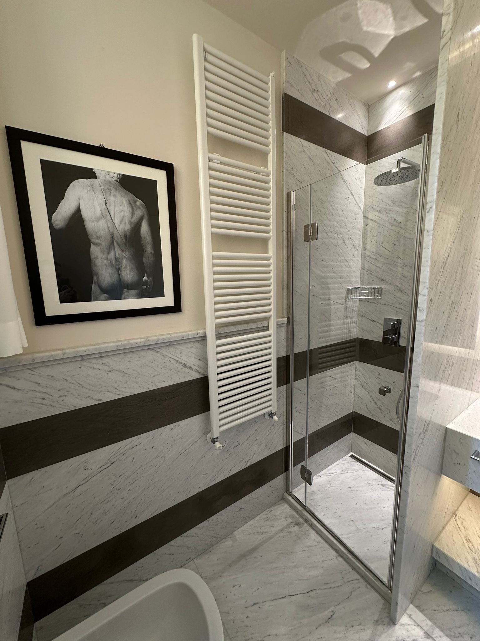 A modern bathroom with a glass shower stall, marble walls, a framed black and white art print of a back view of a person's upper body, and a white heated towel rail.