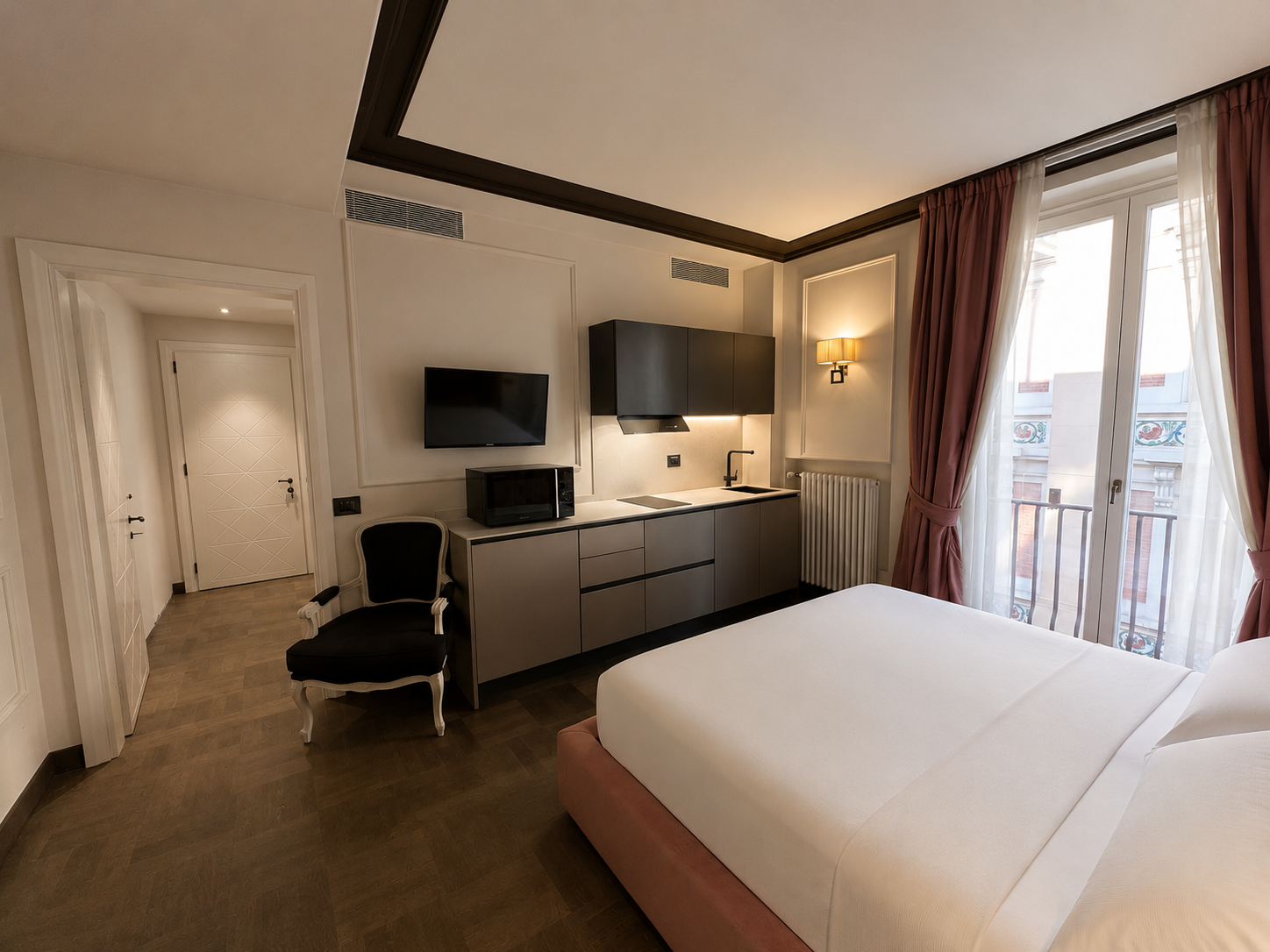 A hotel room with a bed, a black chair, a wall-mounted television, a microwave, black cabinetry, a kitchen sink, a window with pink curtains, and a door.