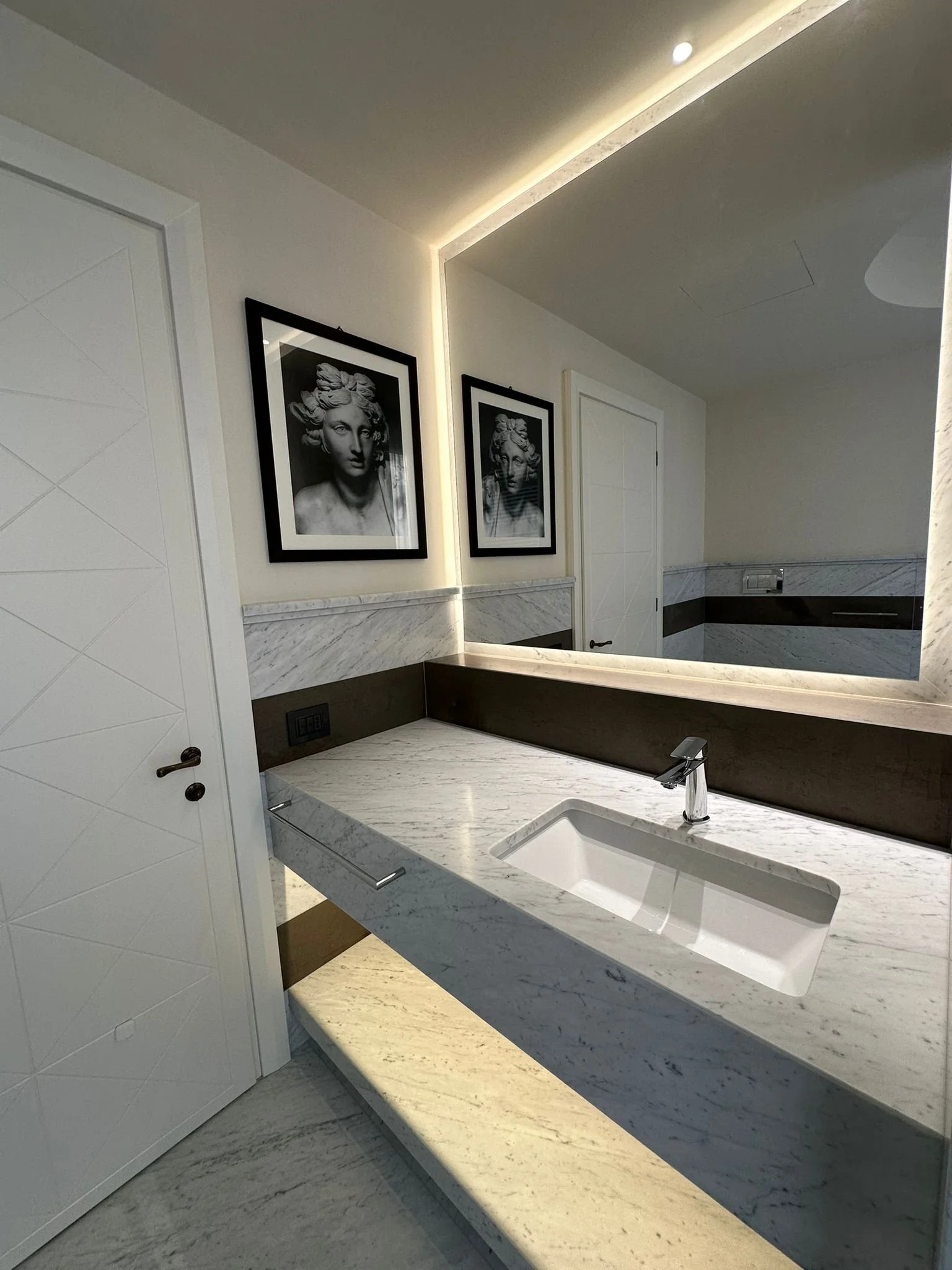 A modern bathroom with white marble countertop, rectangular sink, and a silver faucet. The wall has black and white framed artwork of a woman's face, and the ceiling features warm LED strip lighting. Part of a white door with a dark handle is visible