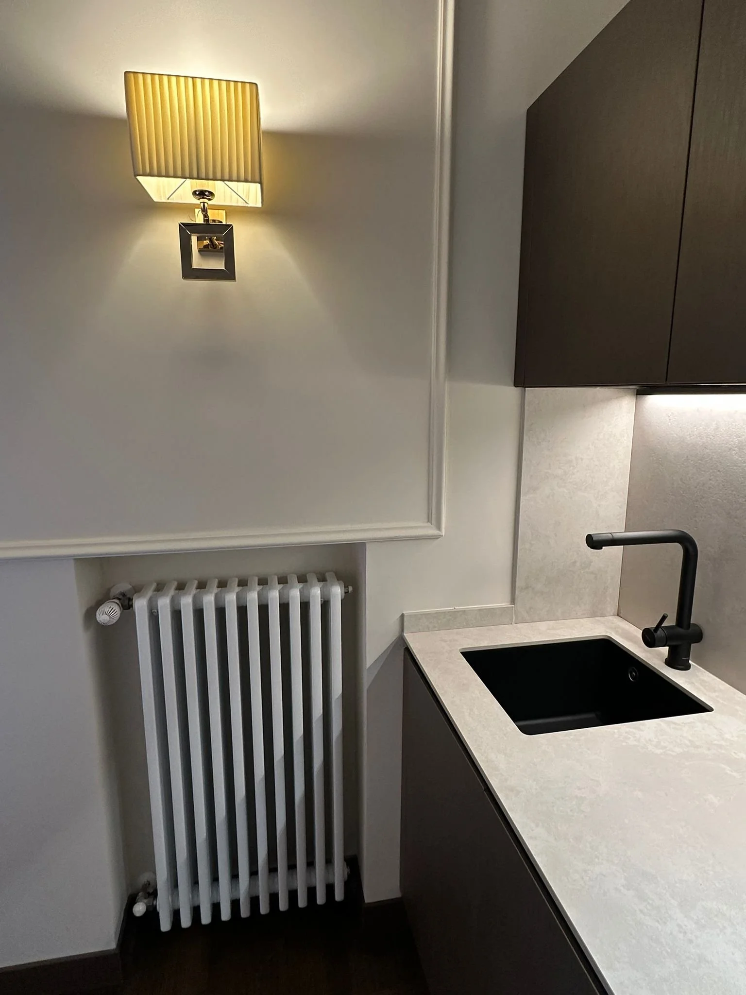 A modern kitchen with a black sink, a black faucet, a beige countertop, dark cabinets, a white radiator, and a wall-mounted lamp with a beige lampshade.