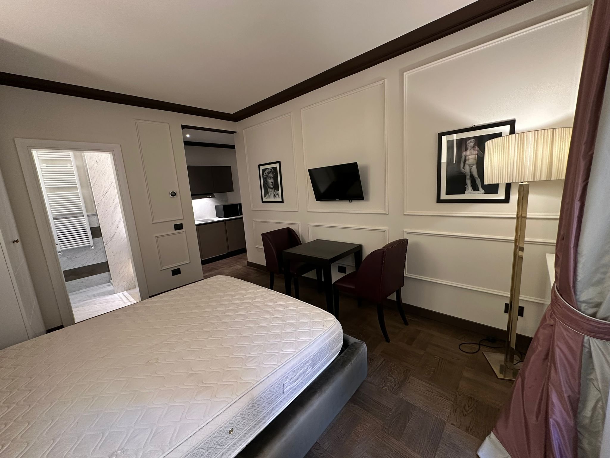 A hotel room with a mattress, a small table with two chairs, framed artwork on the wall, a lamp, and a doorway leading to a bathroom.