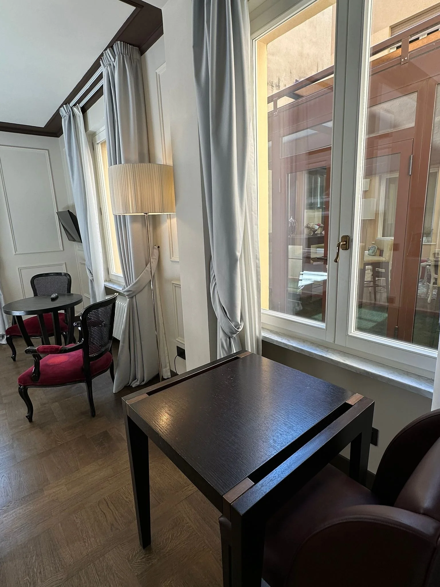Room with a window showing an outdoor balcony, black tables, red velvet chairs, a floor lamp, and white curtains.