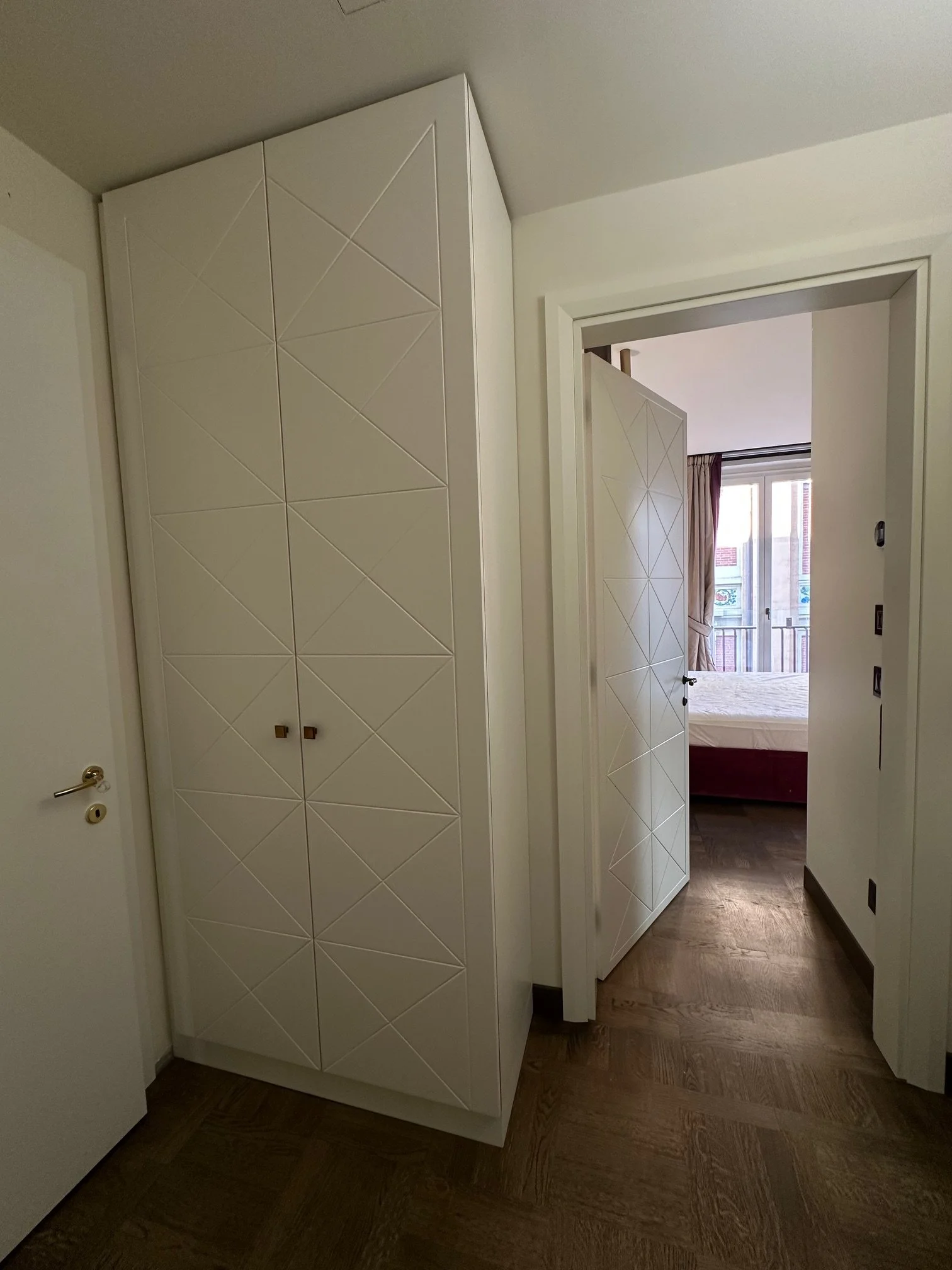 Interior of an apartment with a white built-in wardrobe, an open doorway revealing a bedroom with a window and curtains, dark wooden flooring, and white walls.