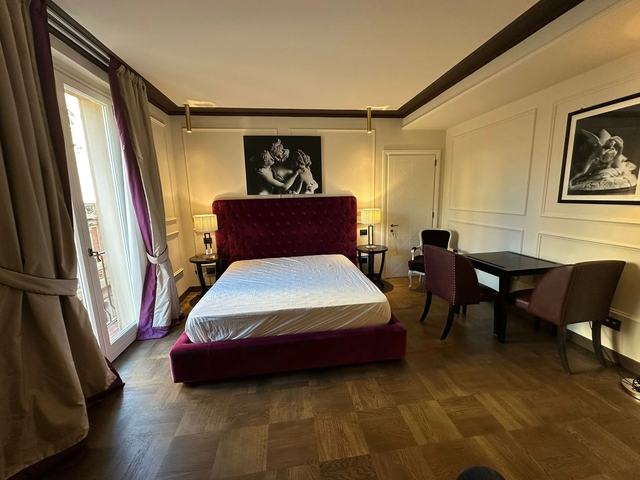 A hotel room with a bed featuring a red headboard, black and white artwork above the bed, beige walls with decorative molding, beige curtains, a window, side tables with lamps, a desk with a chair, and a wooden floor.