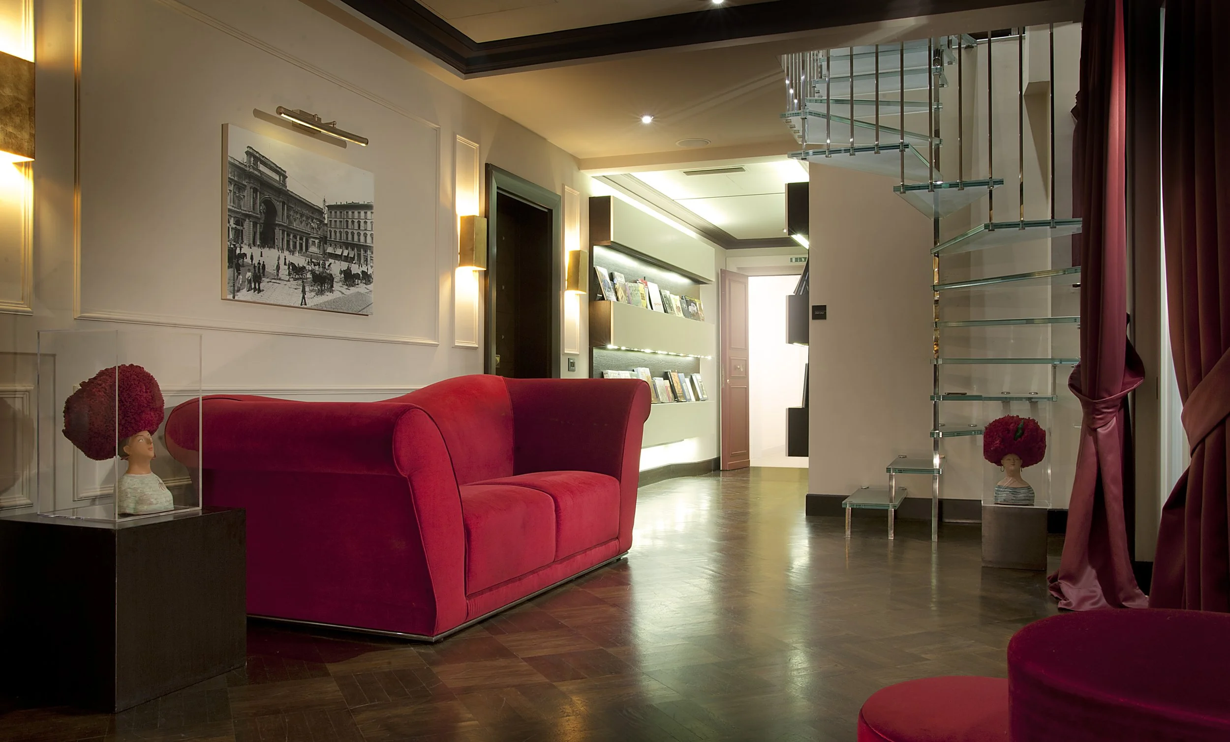 Interior of a modern hotel lobby with a red velvet sofa, decorative mannequins with large red floral headpieces, and a glass staircase. There is a framed black and white photograph on the wall.